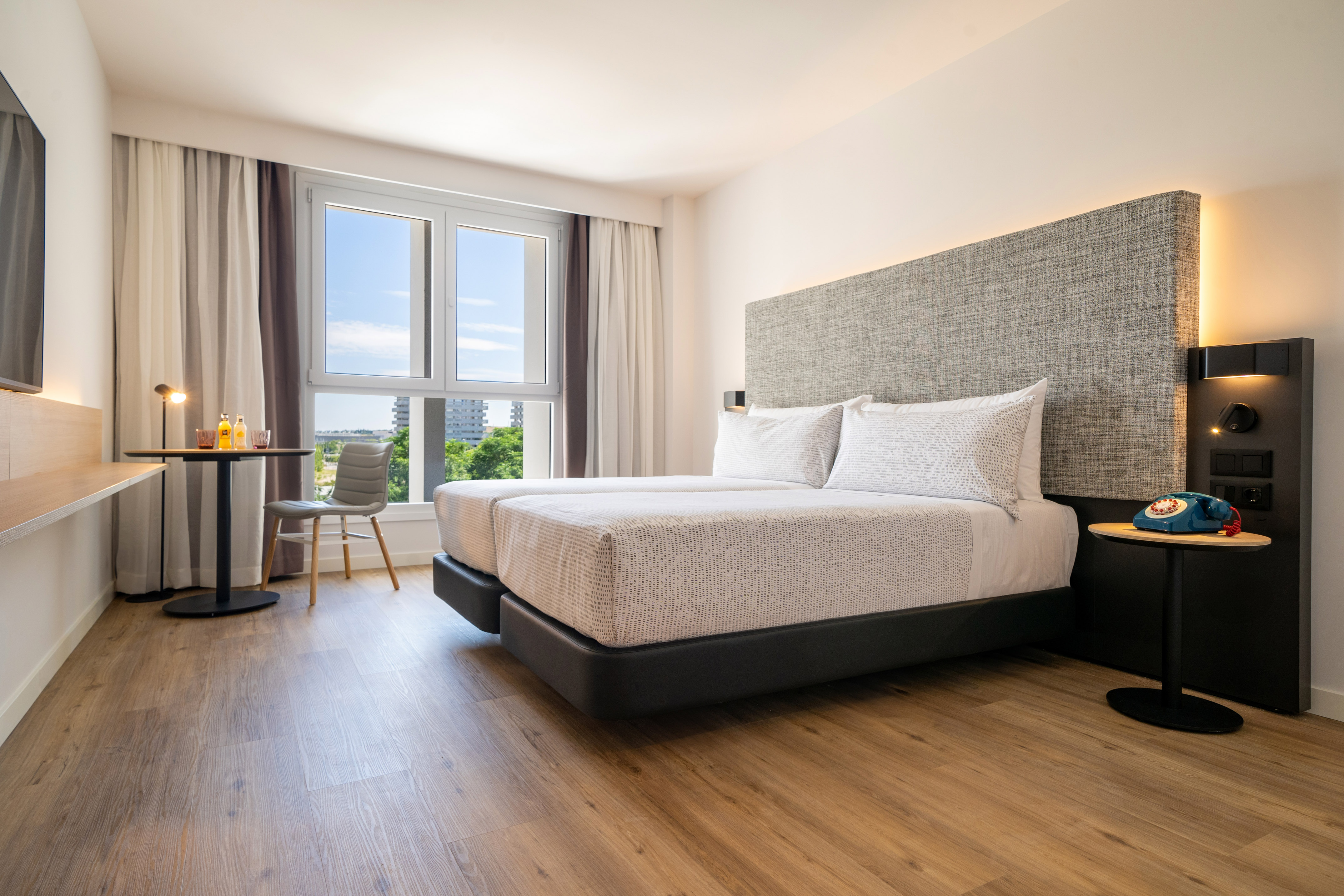 Modern hotel room with twin beds, city view window, and wood floor.