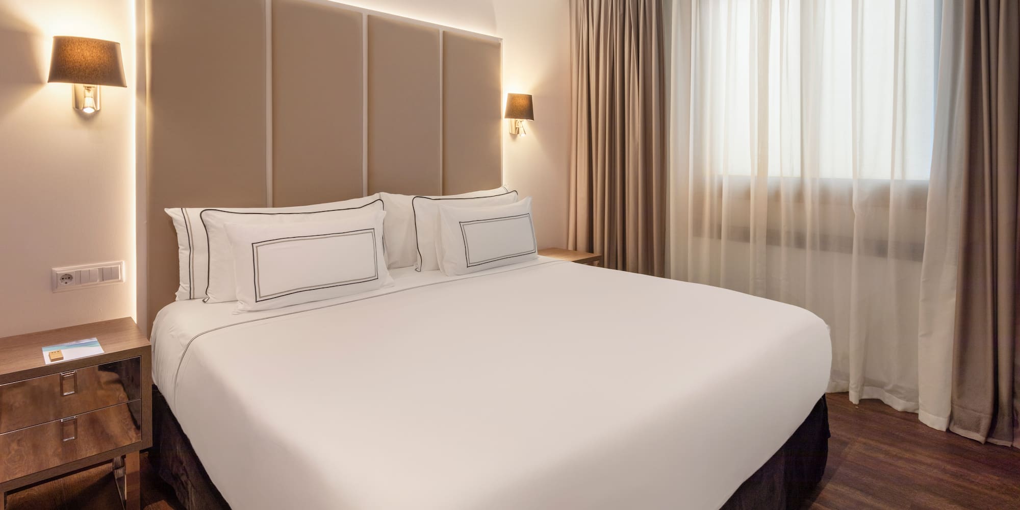 Modern hotel room with a large bed, beige headboard, and warm lighting.