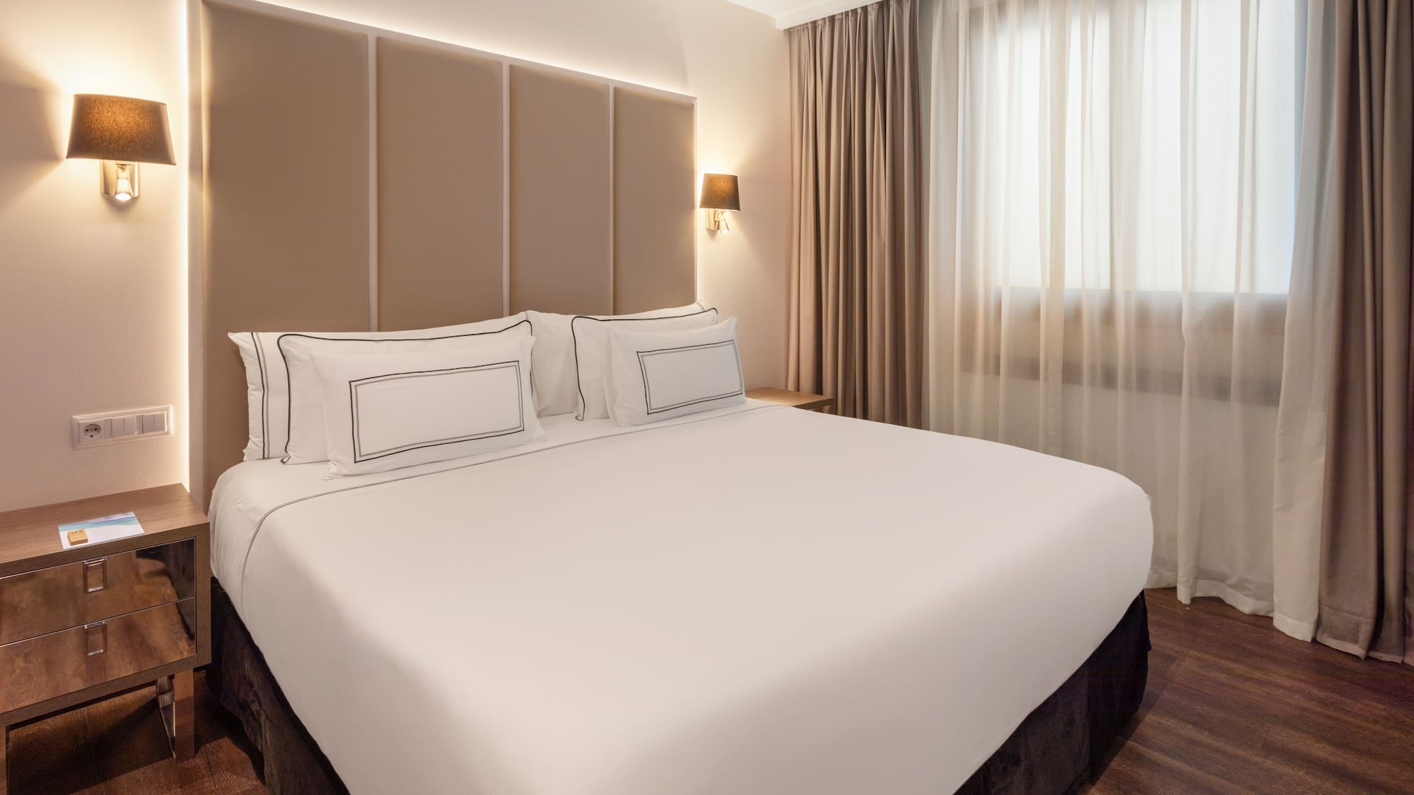 Modern hotel room with a large bed, beige headboard, and warm lighting.