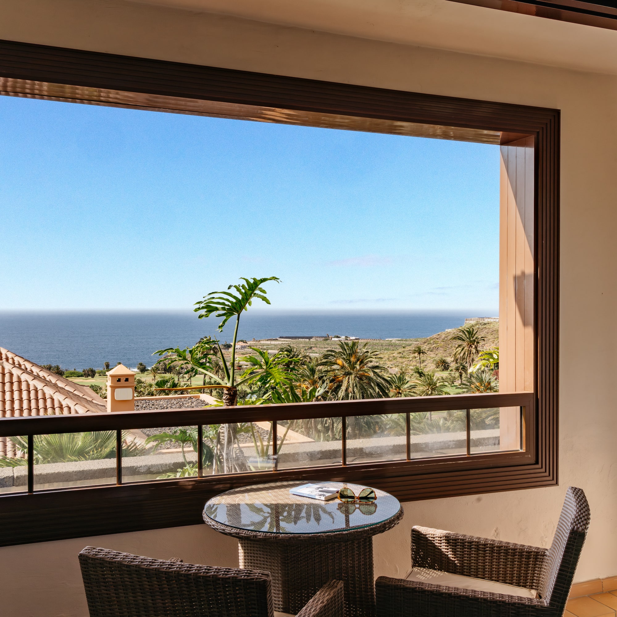 Balcony ocean view with palm trees, blue sky, and wicker furniture.