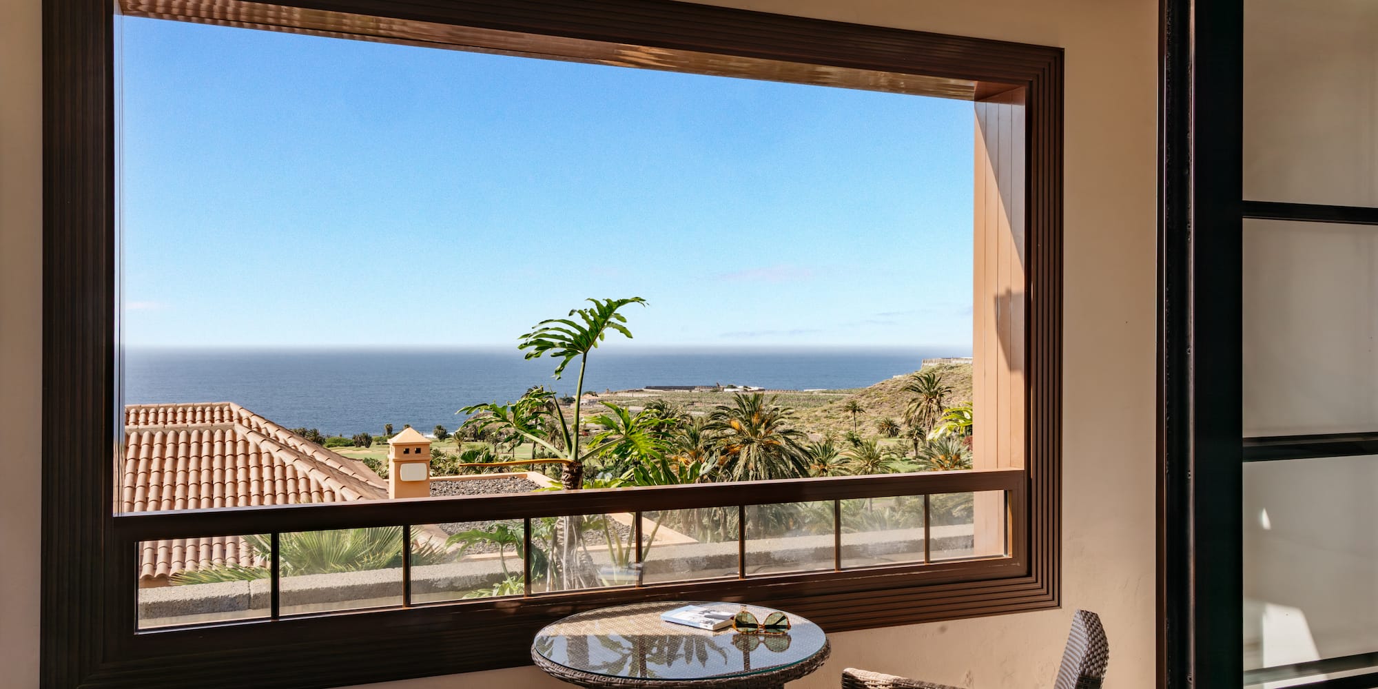 Balcony ocean view with palm trees, blue sky, and wicker furniture.