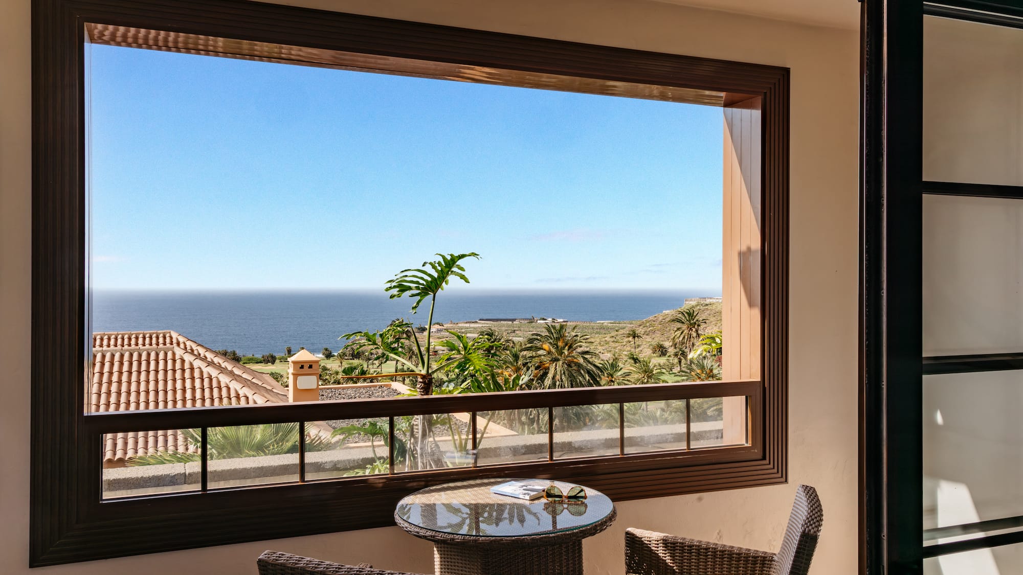 Balcony ocean view with palm trees, blue sky, and wicker furniture.