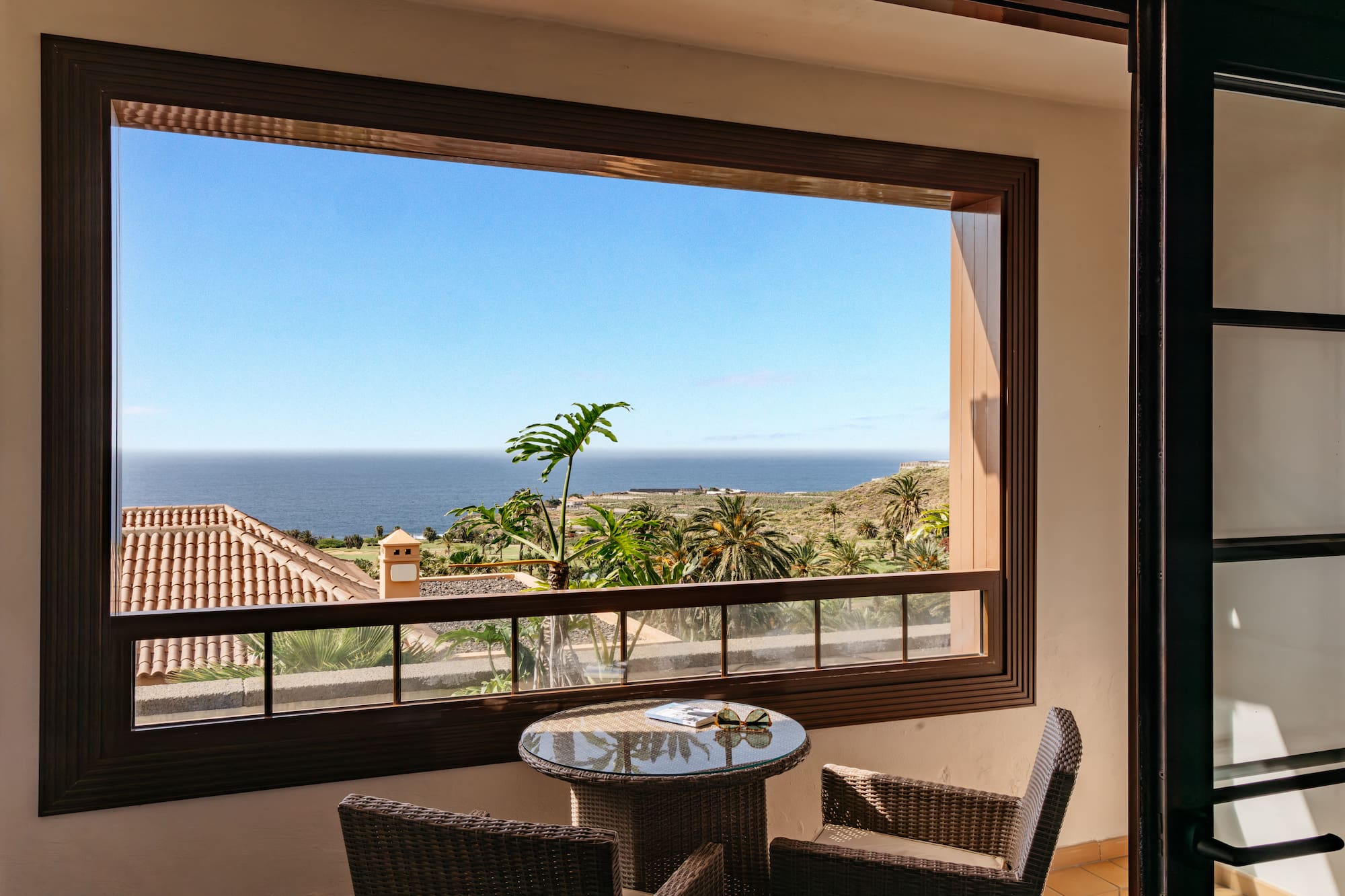 Balcony ocean view with palm trees, blue sky, and wicker furniture.