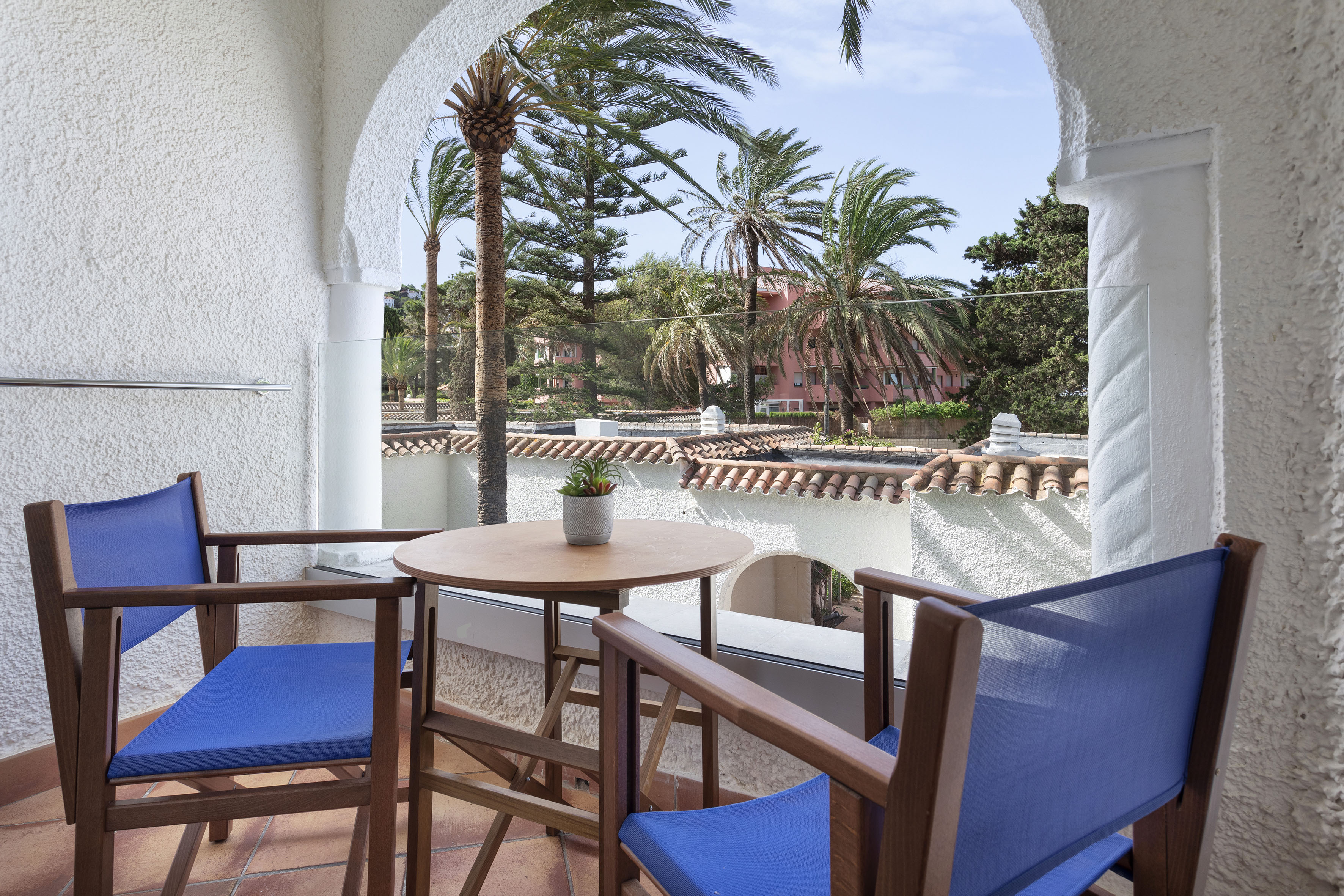 Hotel balcony with blue chairs, small table, and palm tree view.