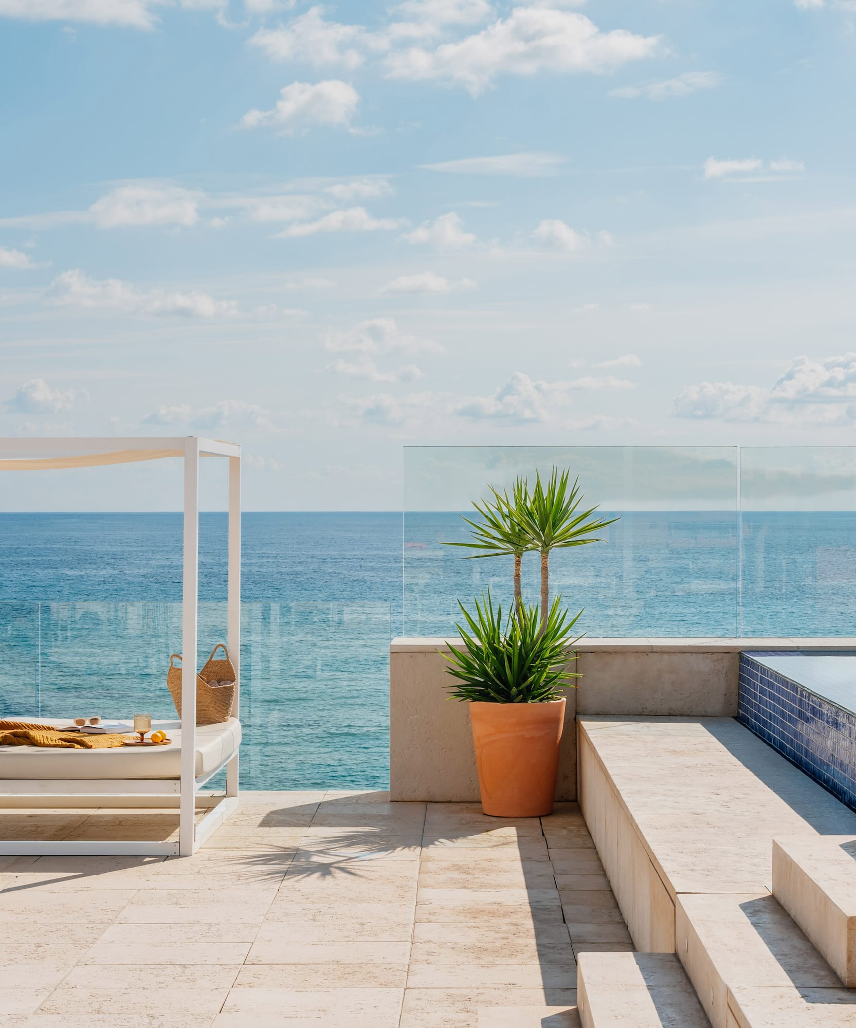 Piscina infinita en terraza con vista al mar, cabaña blanca y planta tropical.