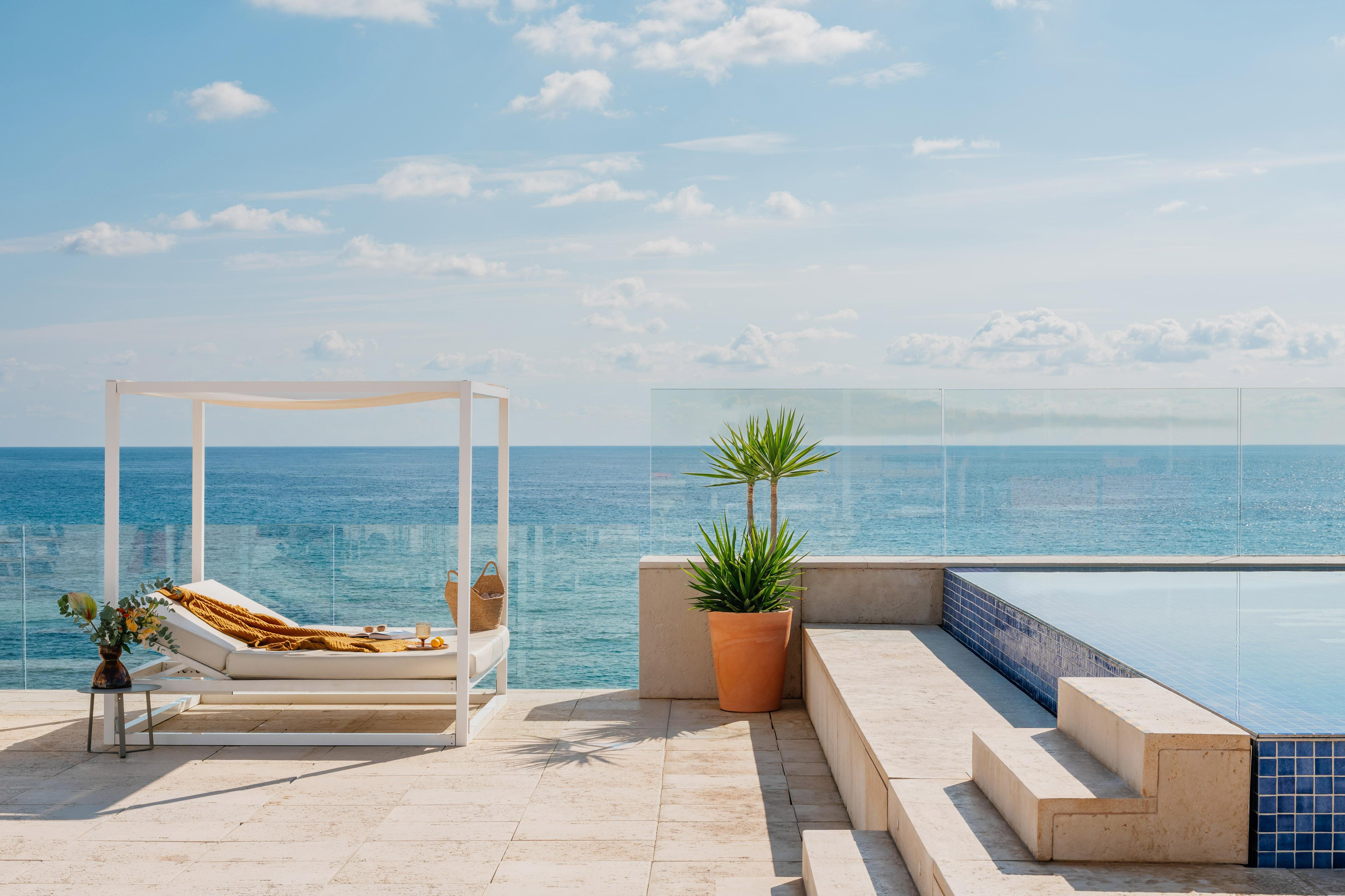 Piscina infinita en terraza con vista al mar, cabaña blanca y planta tropical.