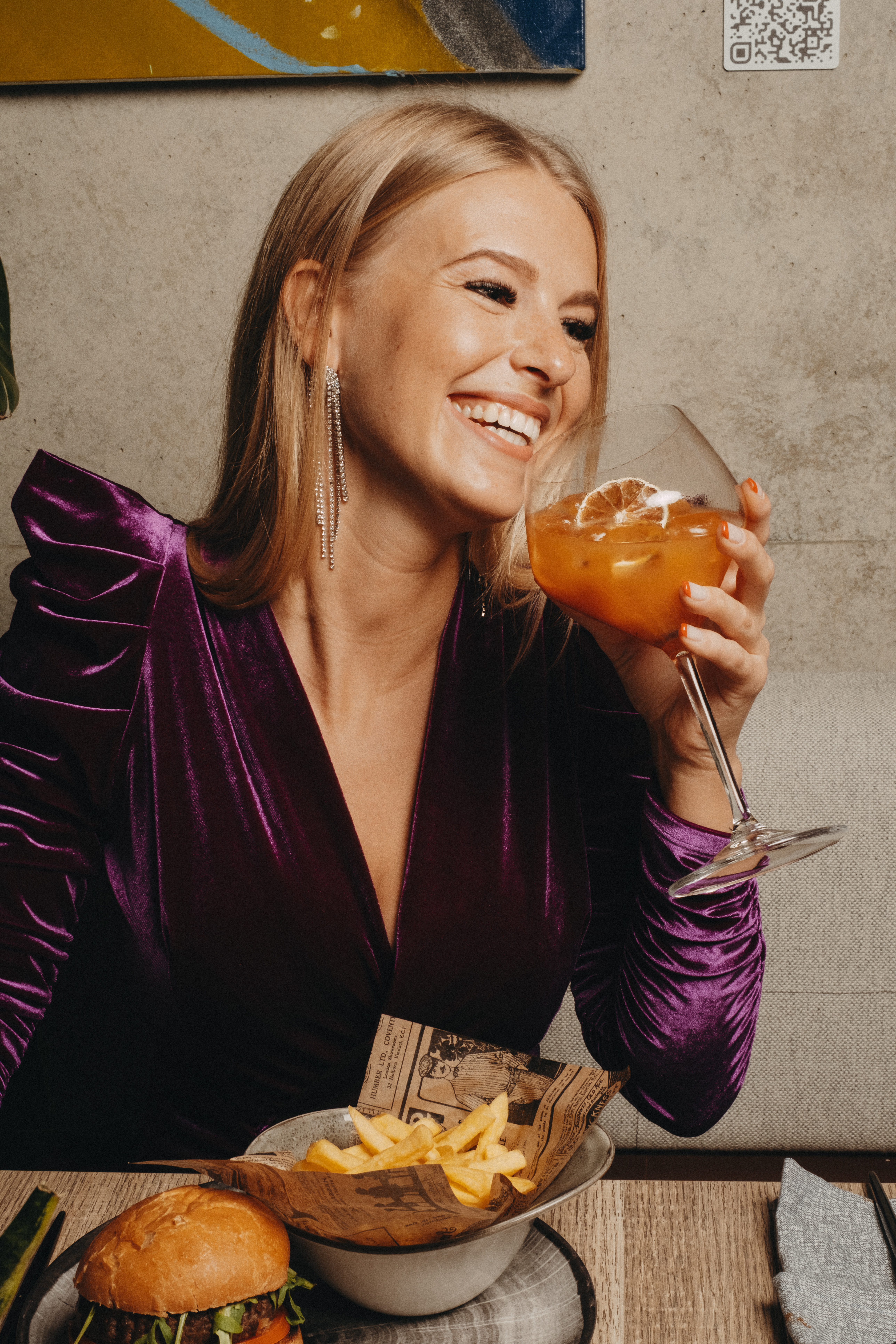 Blonde woman in purple velvet dress smiles, holding cocktail, with burger and fries.
