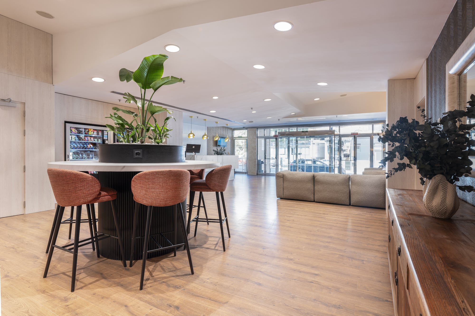 Modern lobby with communal table, orange bar stools, green plants, wood floor.