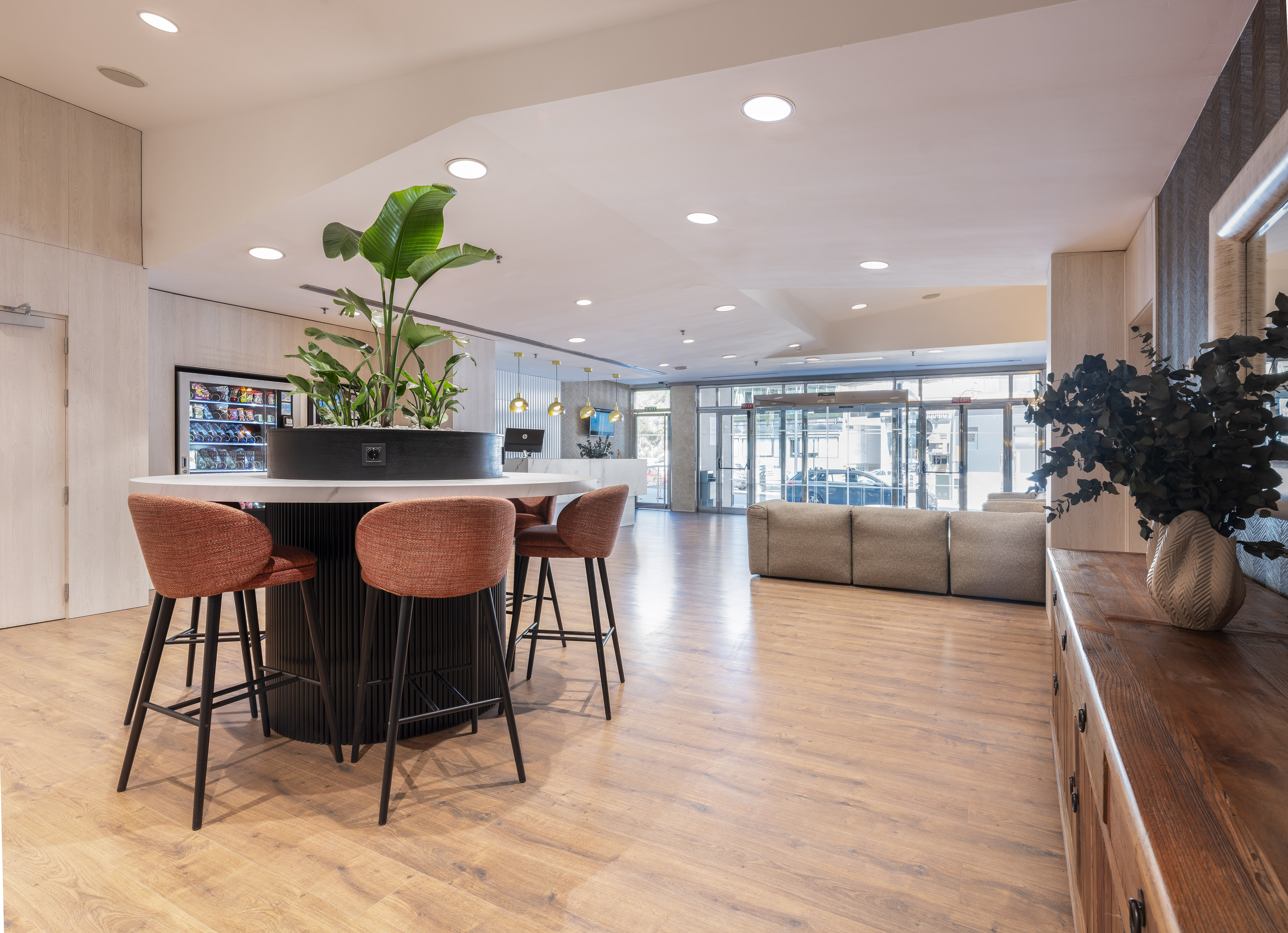 Modern lobby with communal table, orange bar stools, green plants, wood floor.