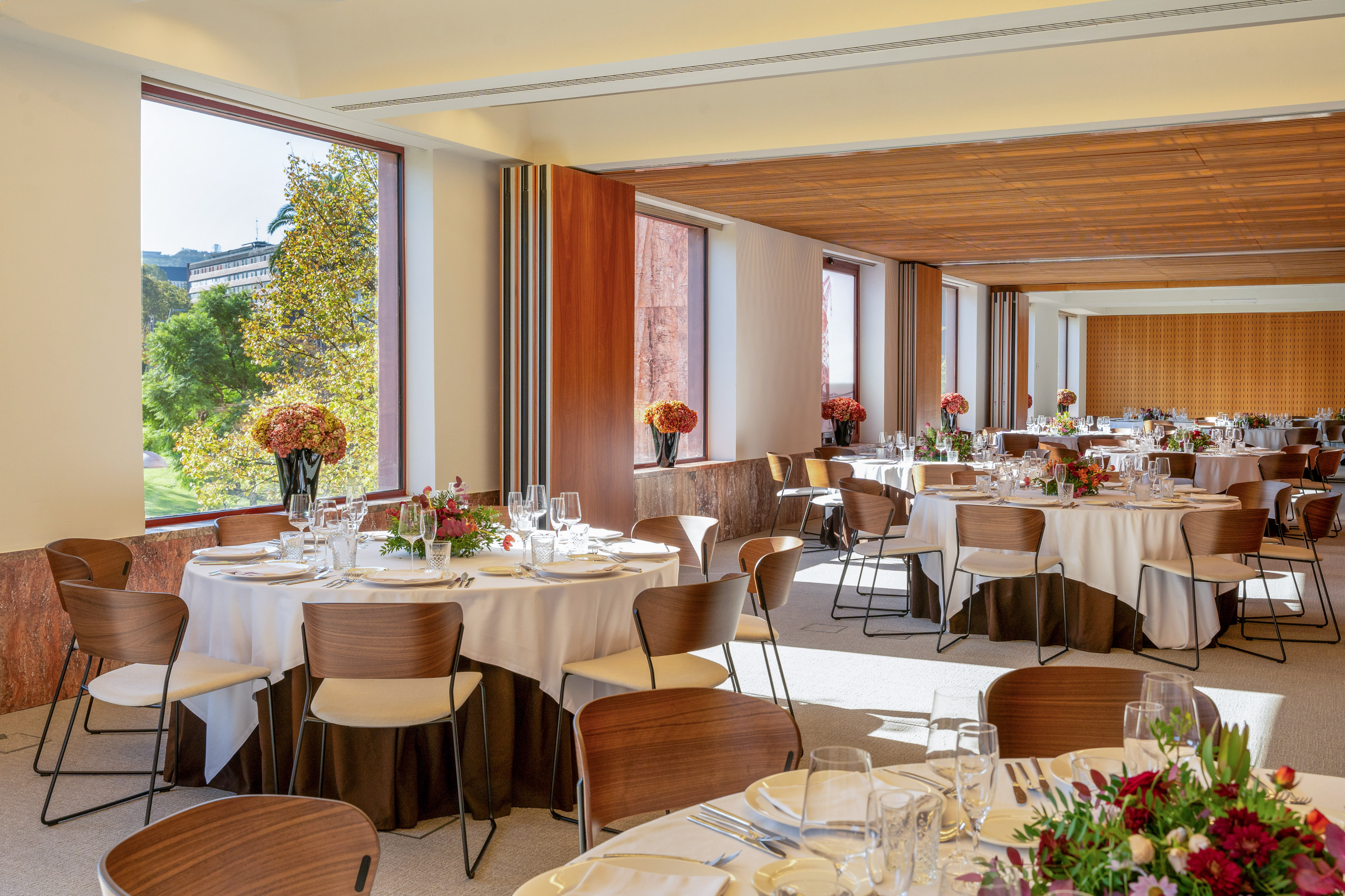 Spacious event hall with set dining tables, modern chairs, and park views.