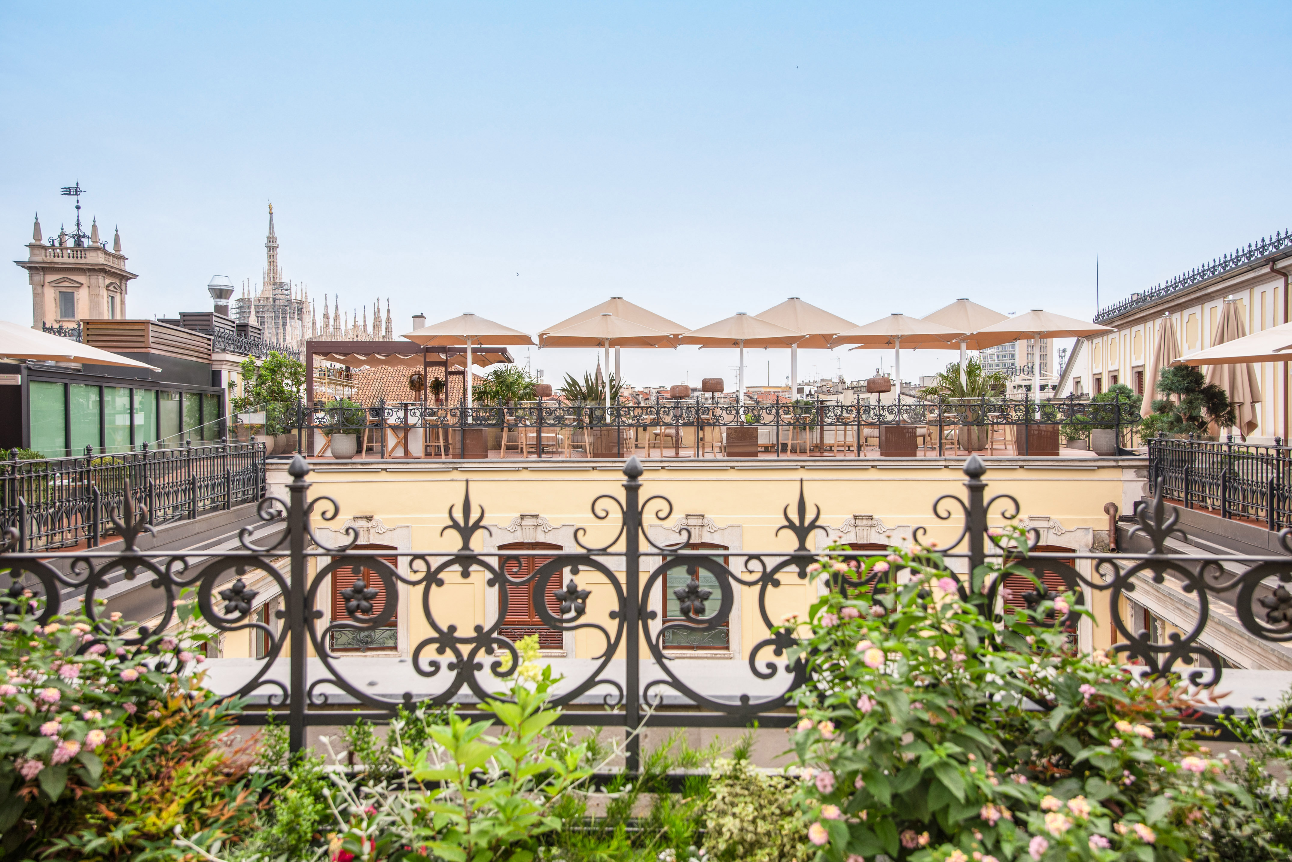 Milan rooftop terrace with Duomo view, sun umbrellas, and ornate railing.