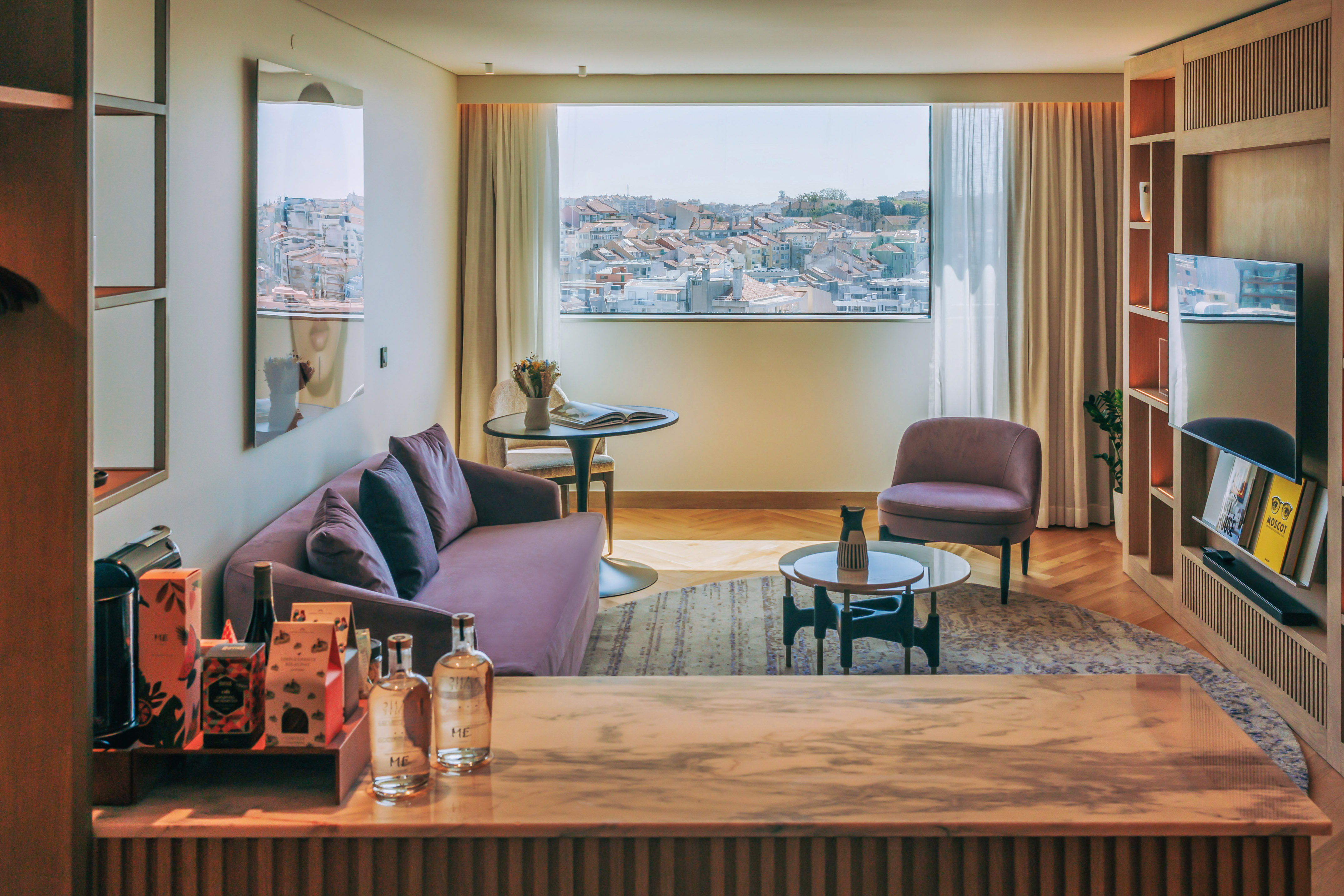 Modern hotel suite living room with city view, purple sofa, and minibar.