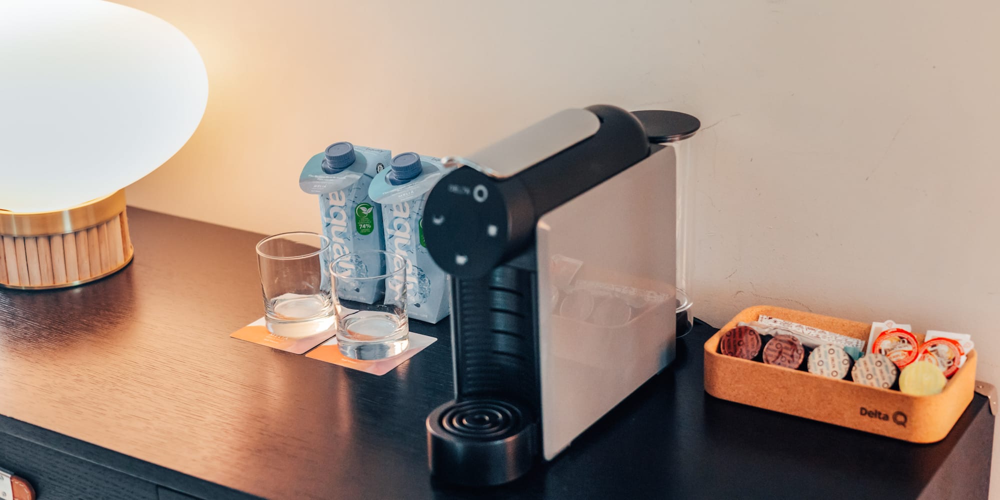 Hotel coffee station: Delta Q machine, water, capsules, and lamp on dark wood.