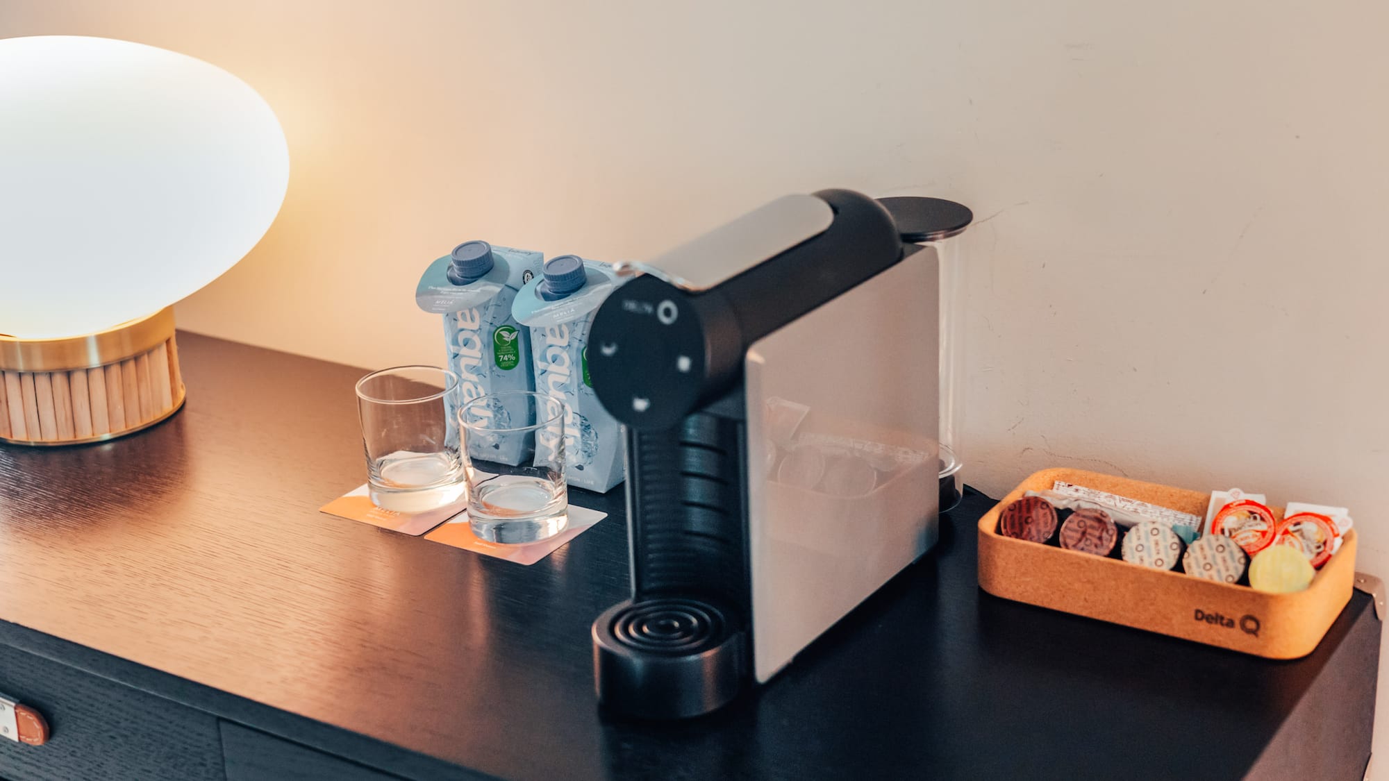 Hotel coffee station: Delta Q machine, water, capsules, and lamp on dark wood.