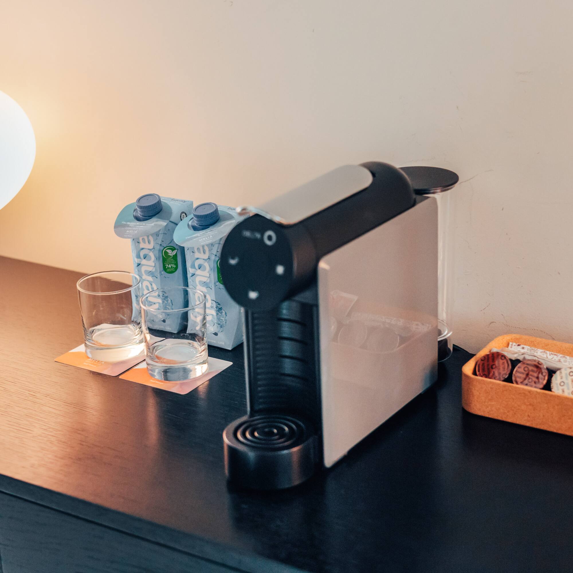 Hotel coffee station: Delta Q machine, water, capsules, and lamp on dark wood.