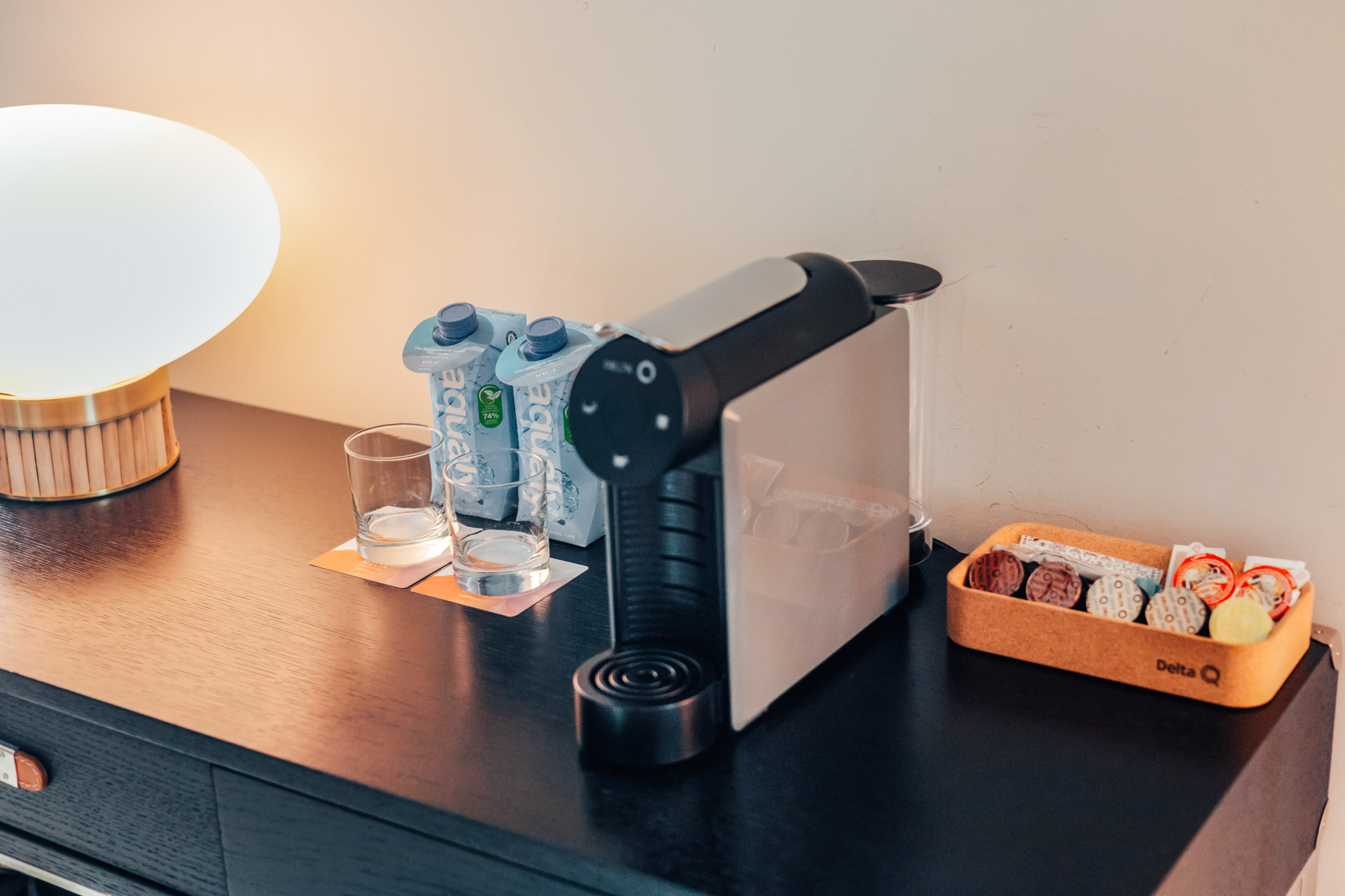 Hotel coffee station: Delta Q machine, water, capsules, and lamp on dark wood.