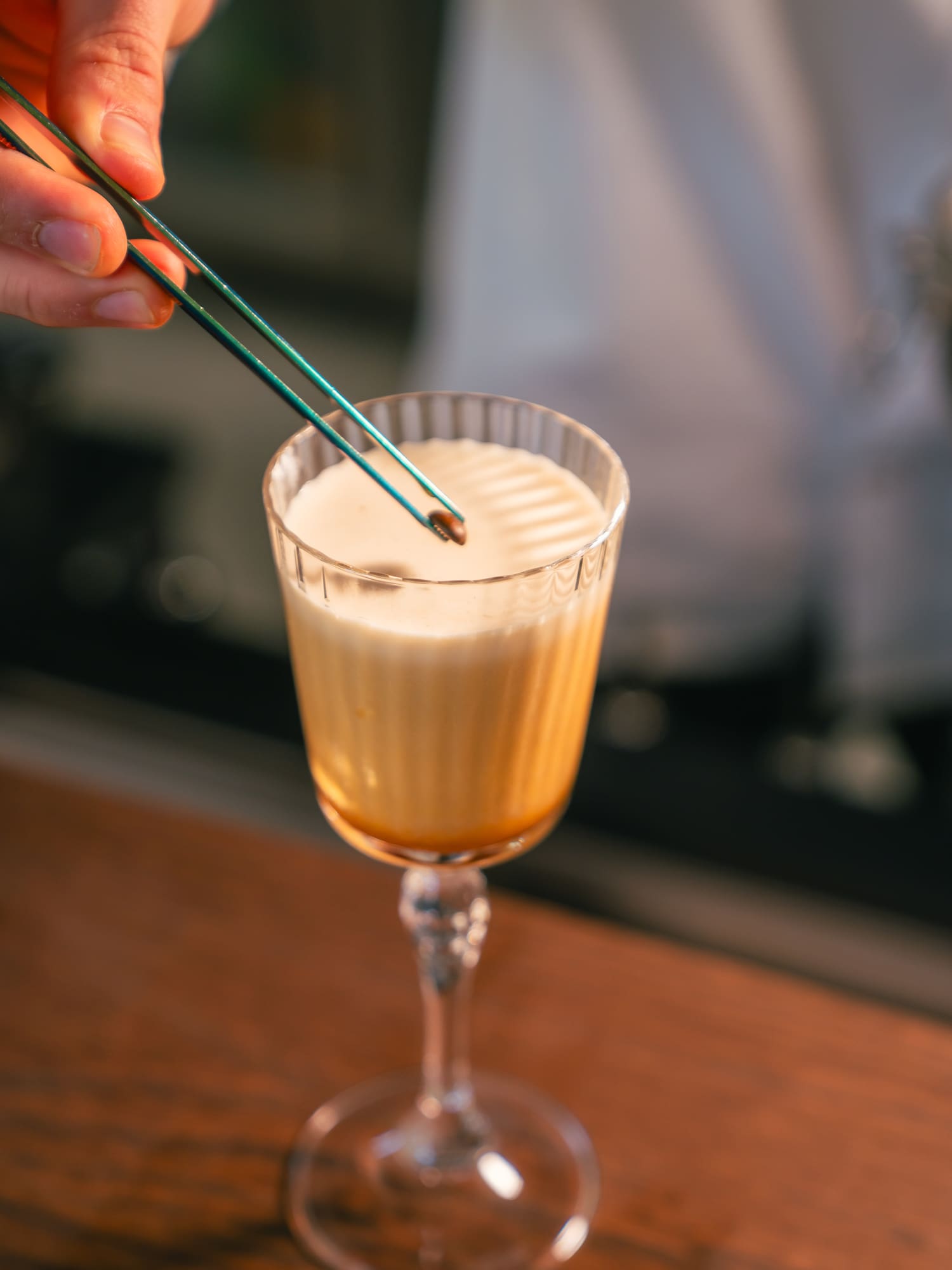 Hand garnishing a creamy coffee cocktail with a coffee bean using tweezers.