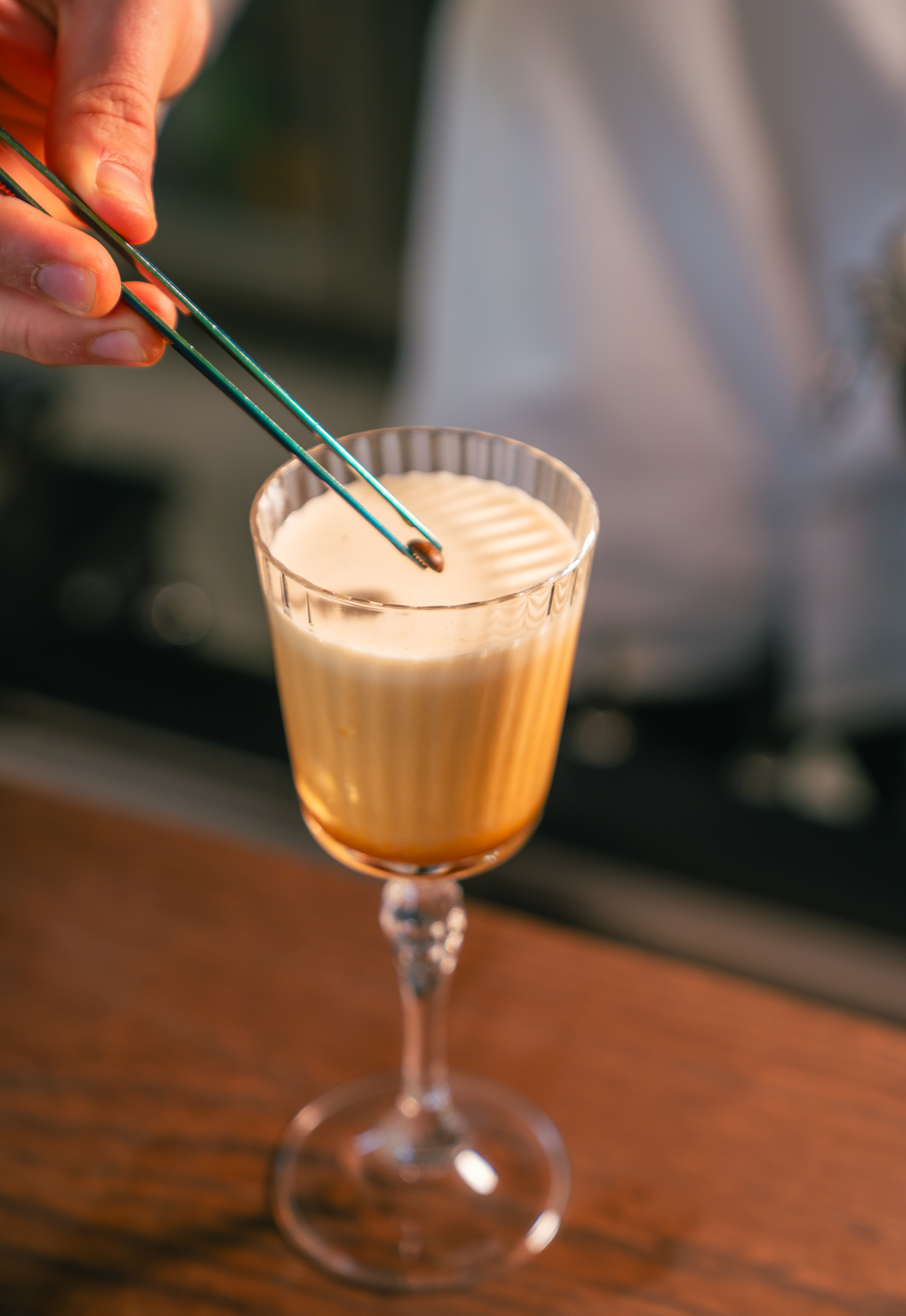 Hand garnishing a creamy coffee cocktail with a coffee bean using tweezers.