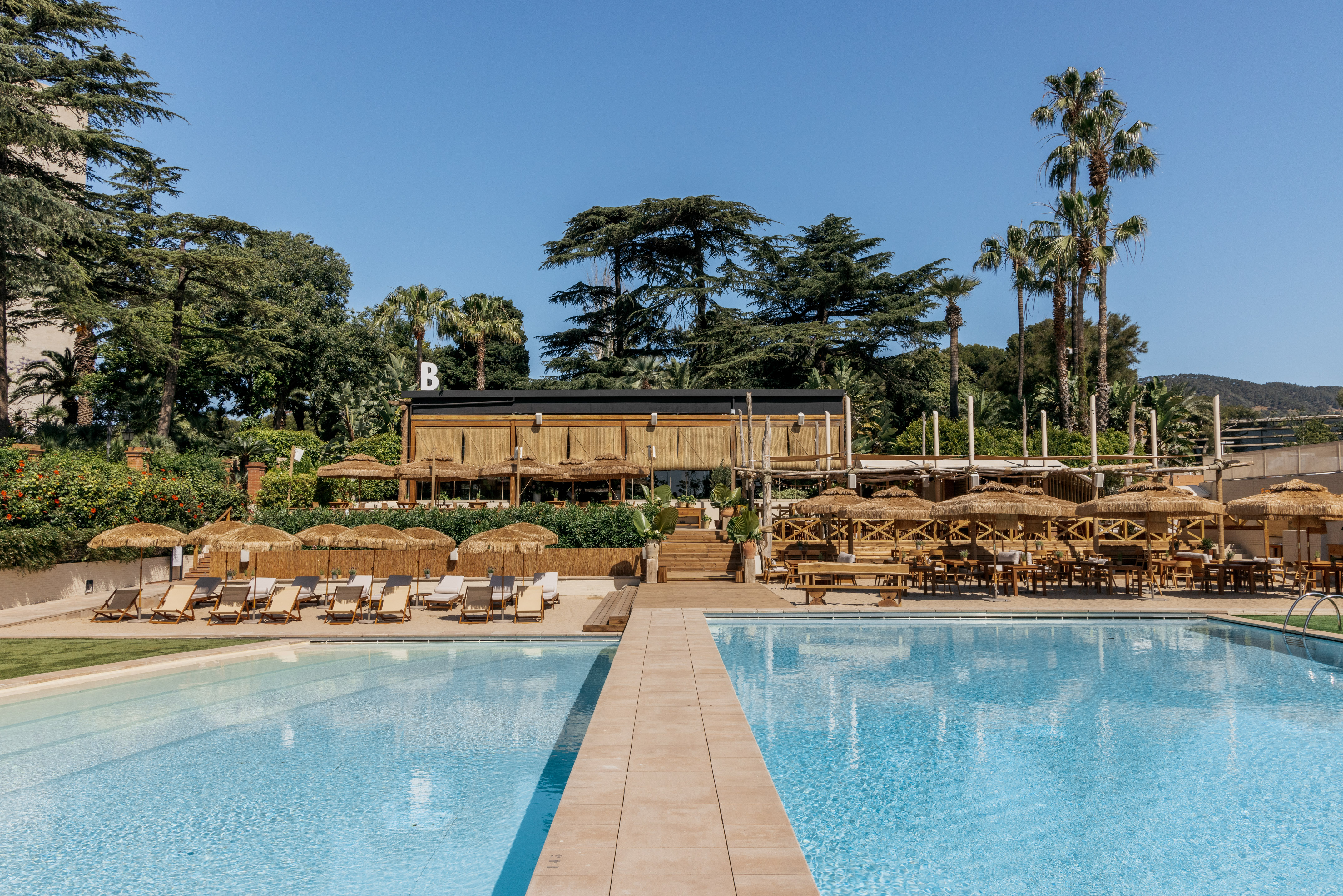Resort pool with B building, sun loungers, straw umbrellas, outdoor dining.