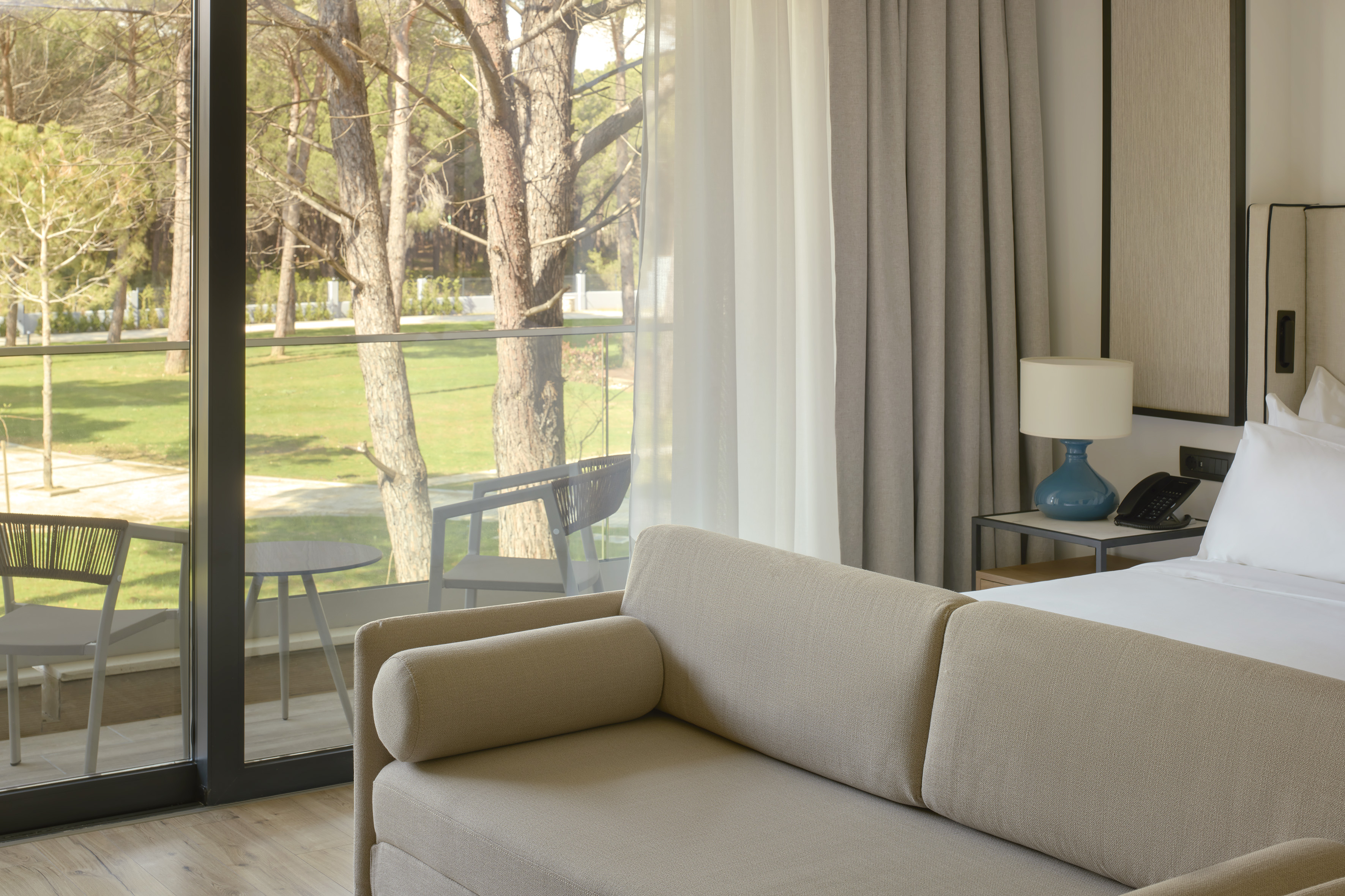 Bright hotel room with sofa, balcony, and lush tree-filled garden view.
