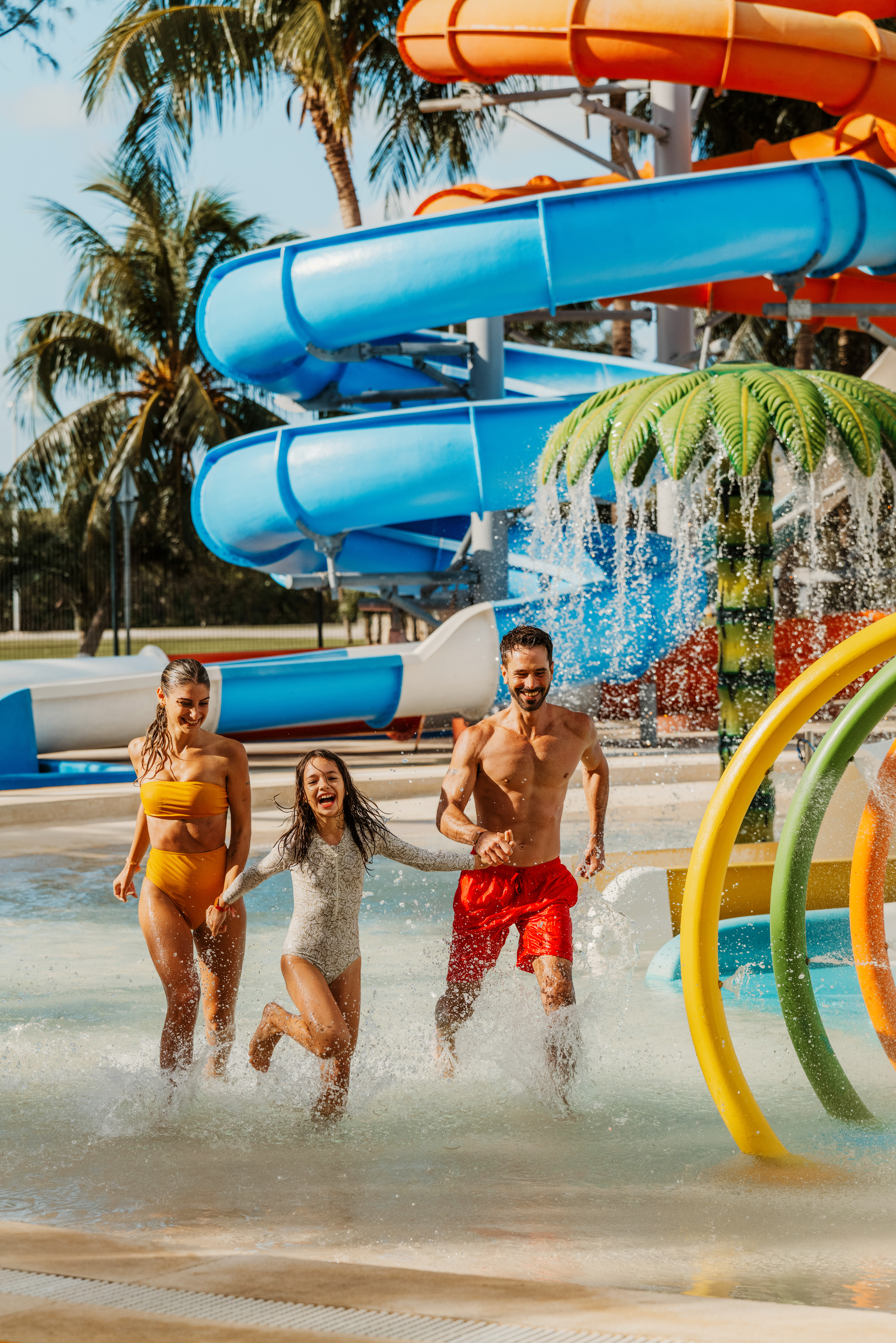 Happy family running in a water park splash pad with colorful slides and palm trees.