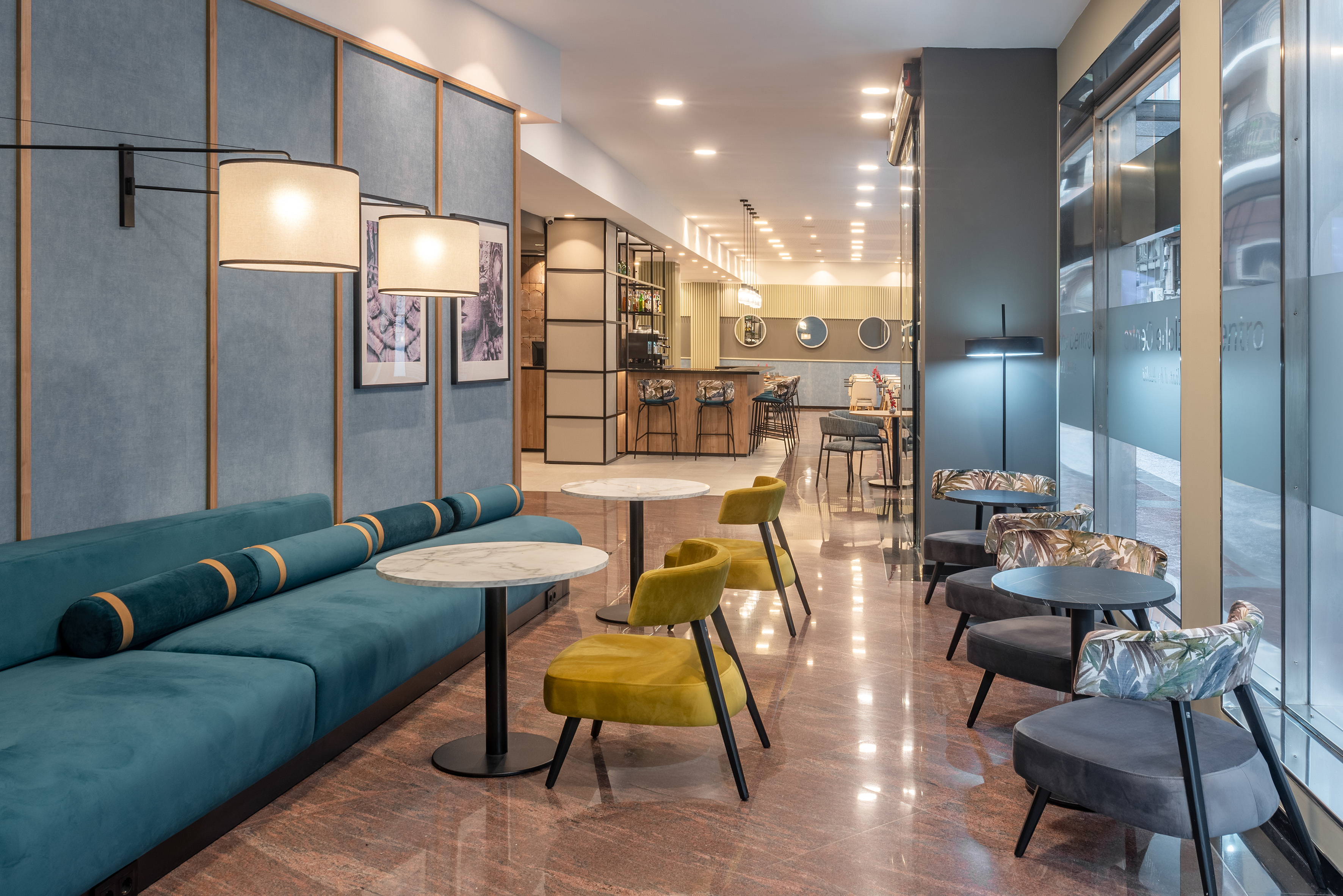 Modern hotel lobby lounge with teal sofa, yellow chairs, marble tables, and bar.