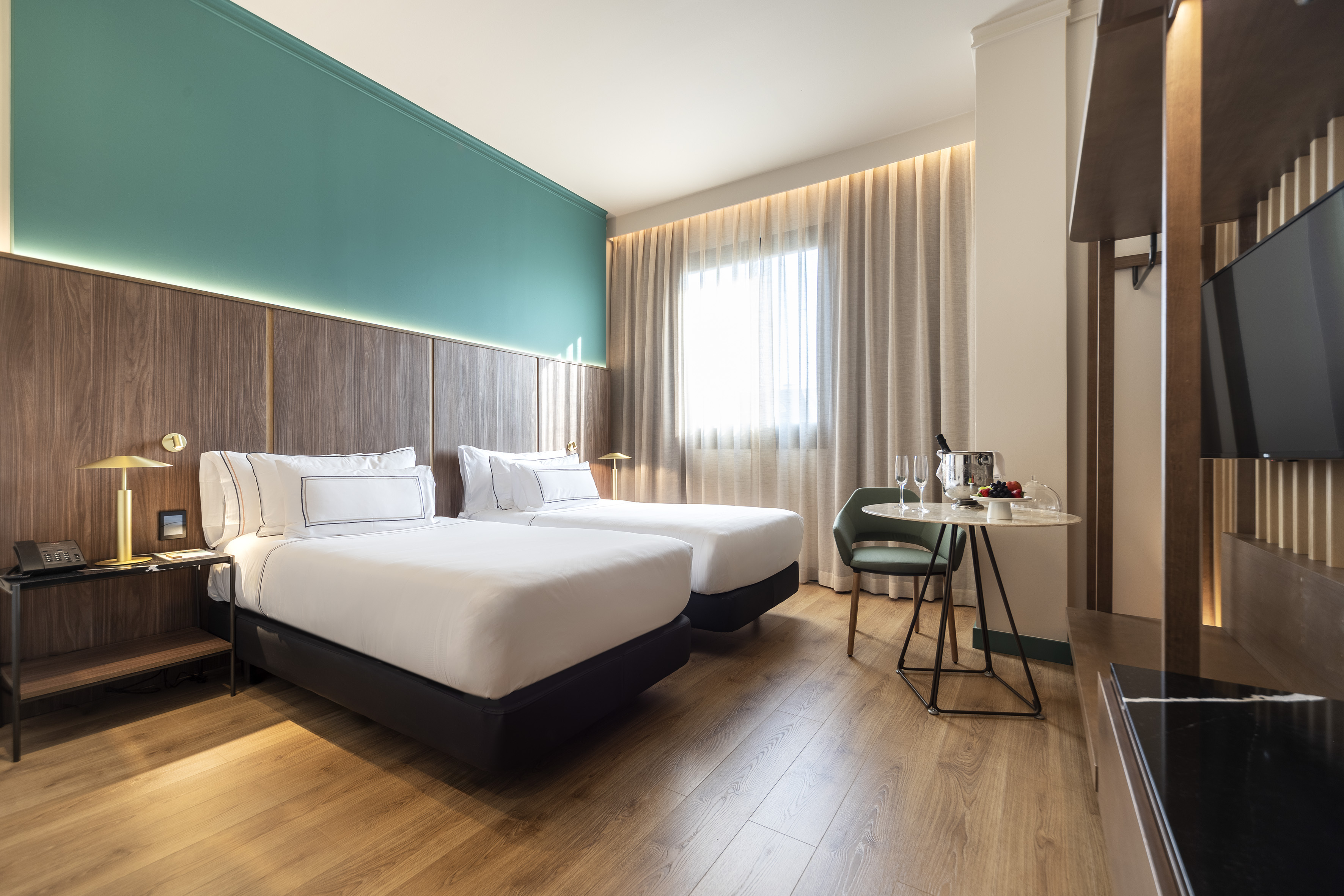 Contemporary hotel room with twin beds, teal wall, wood, and amenities.