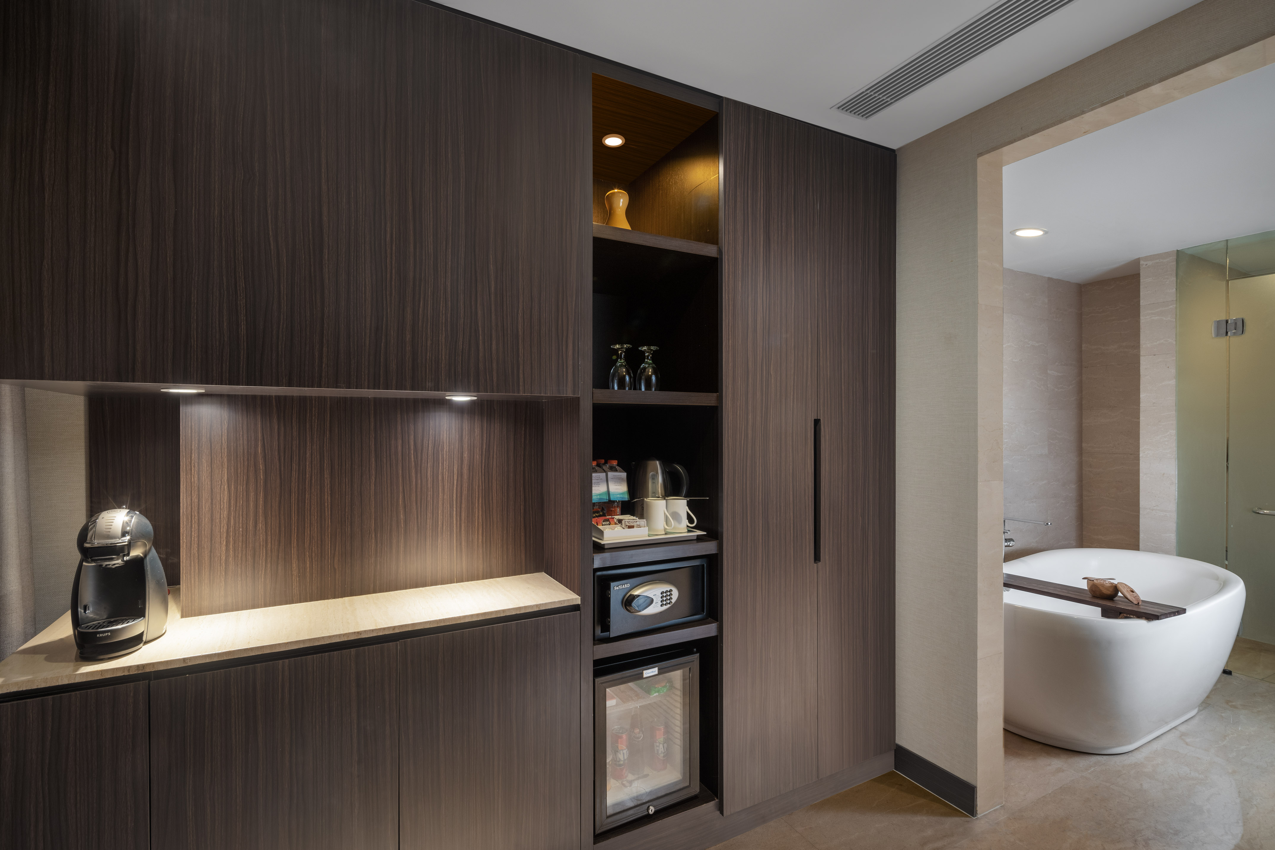 Modern hotel room: dark wood minibar, coffee maker, safe, and luxury bathtub.