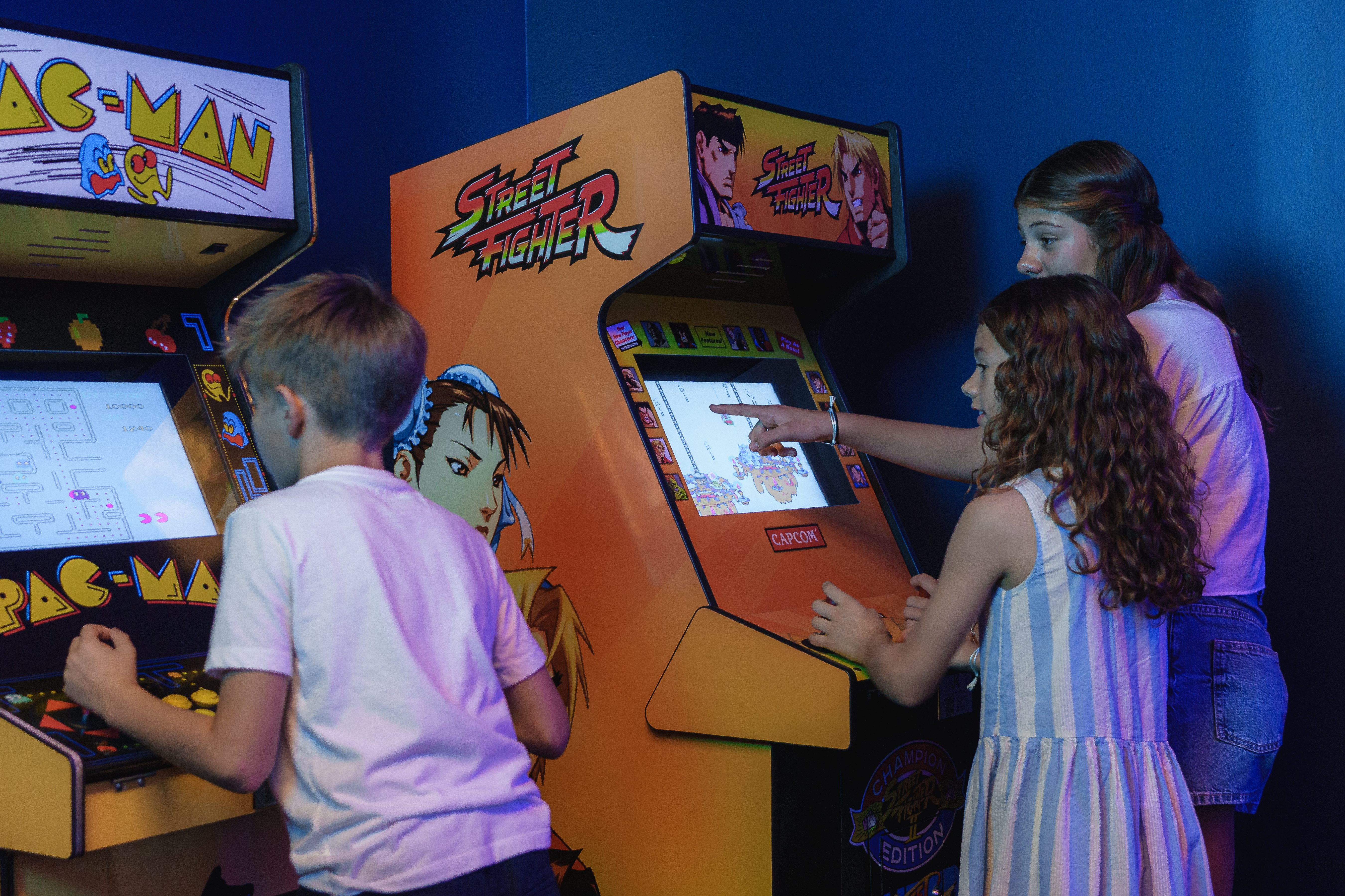 Children playing classic arcade games: boy on Pac-Man, two girls on Street Fighter.