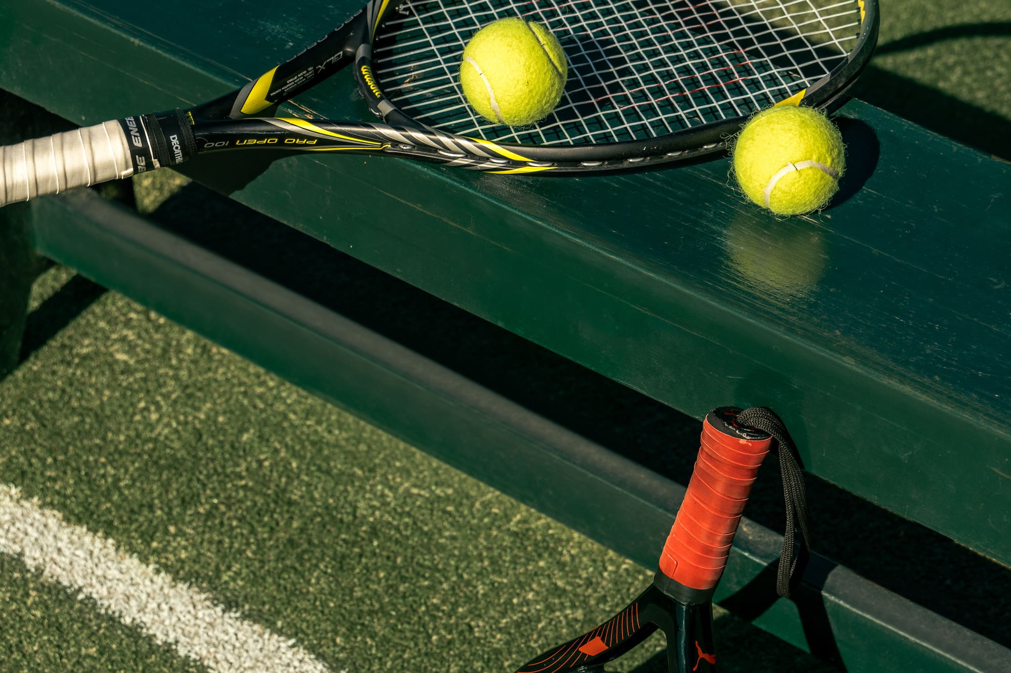 Tennis racket, two balls on bench; padel racket, ball on green court.