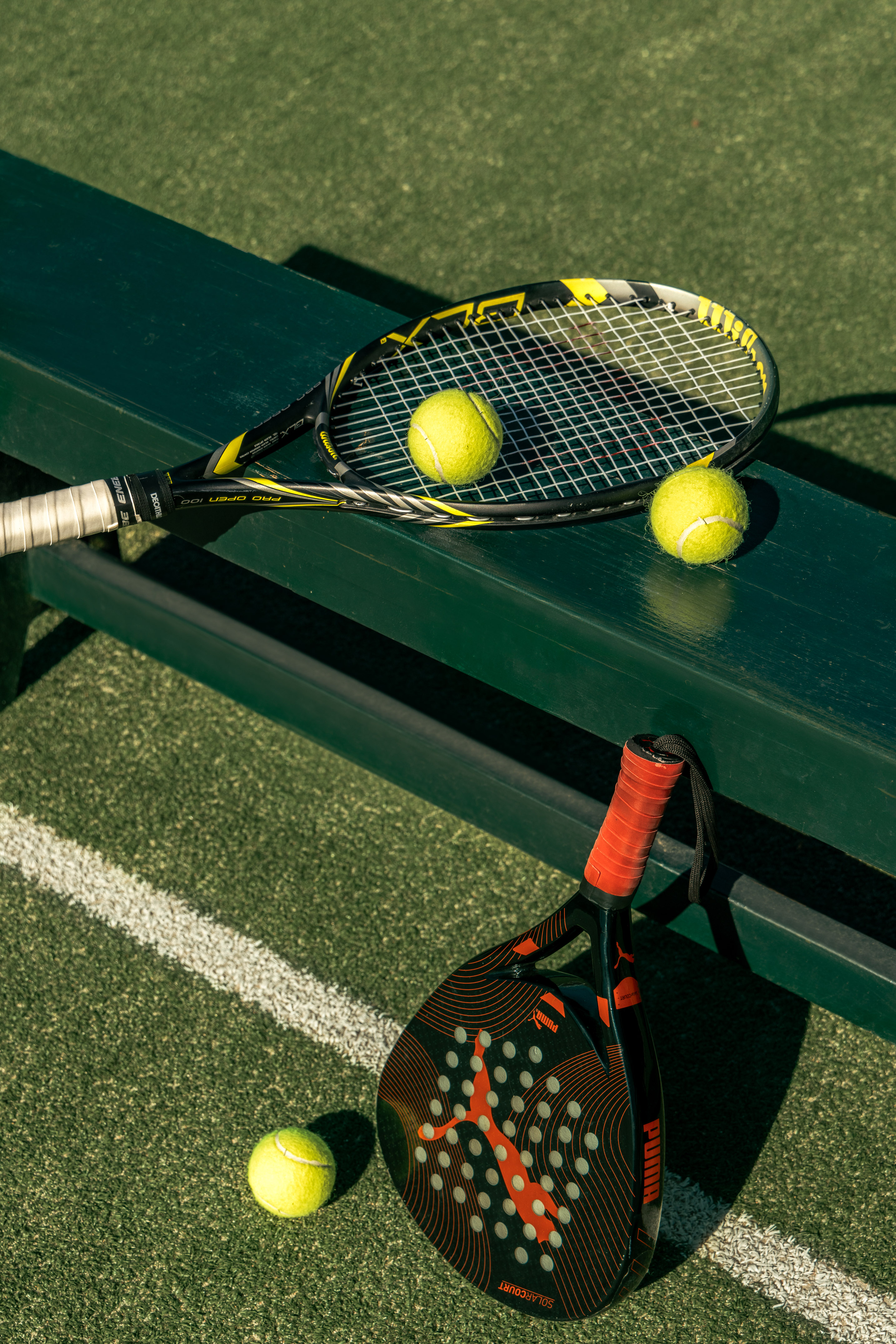 Tennis racket, two balls on bench; padel racket, ball on green court.