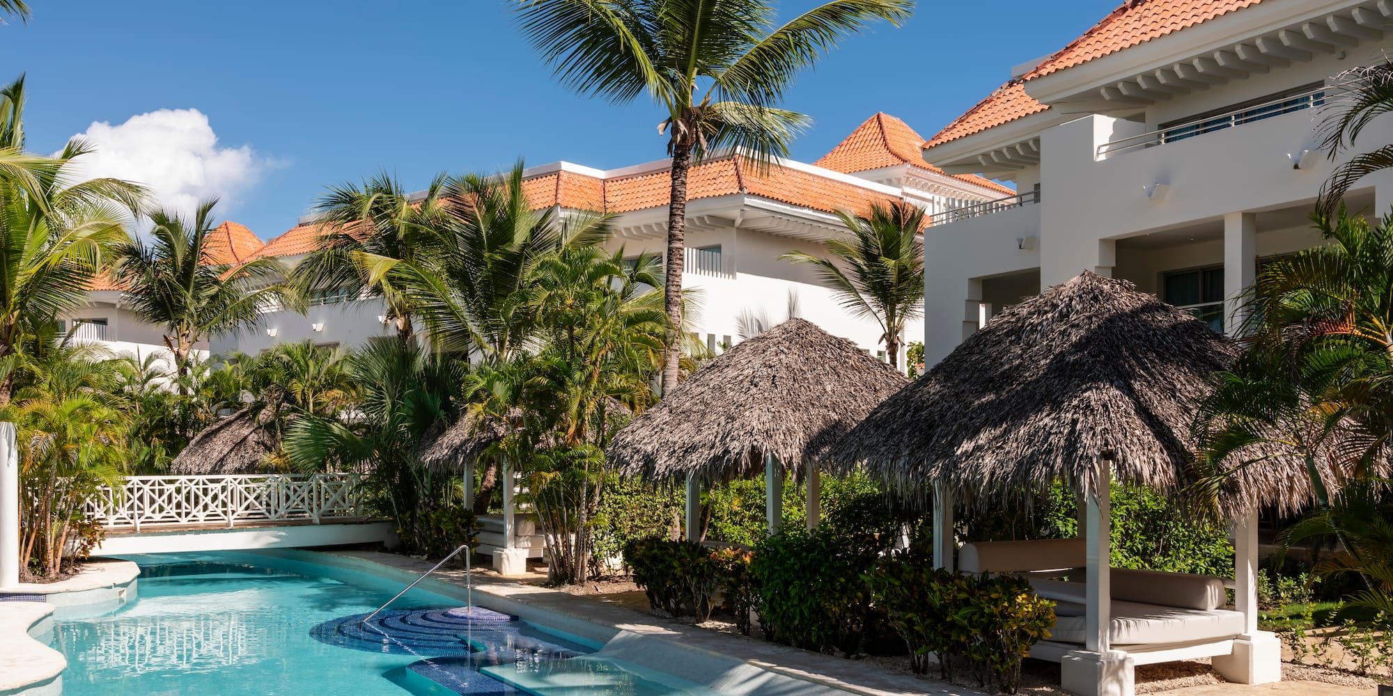 Sunny tropical resort pool with white buildings, palm trees, and thatched cabanas.
