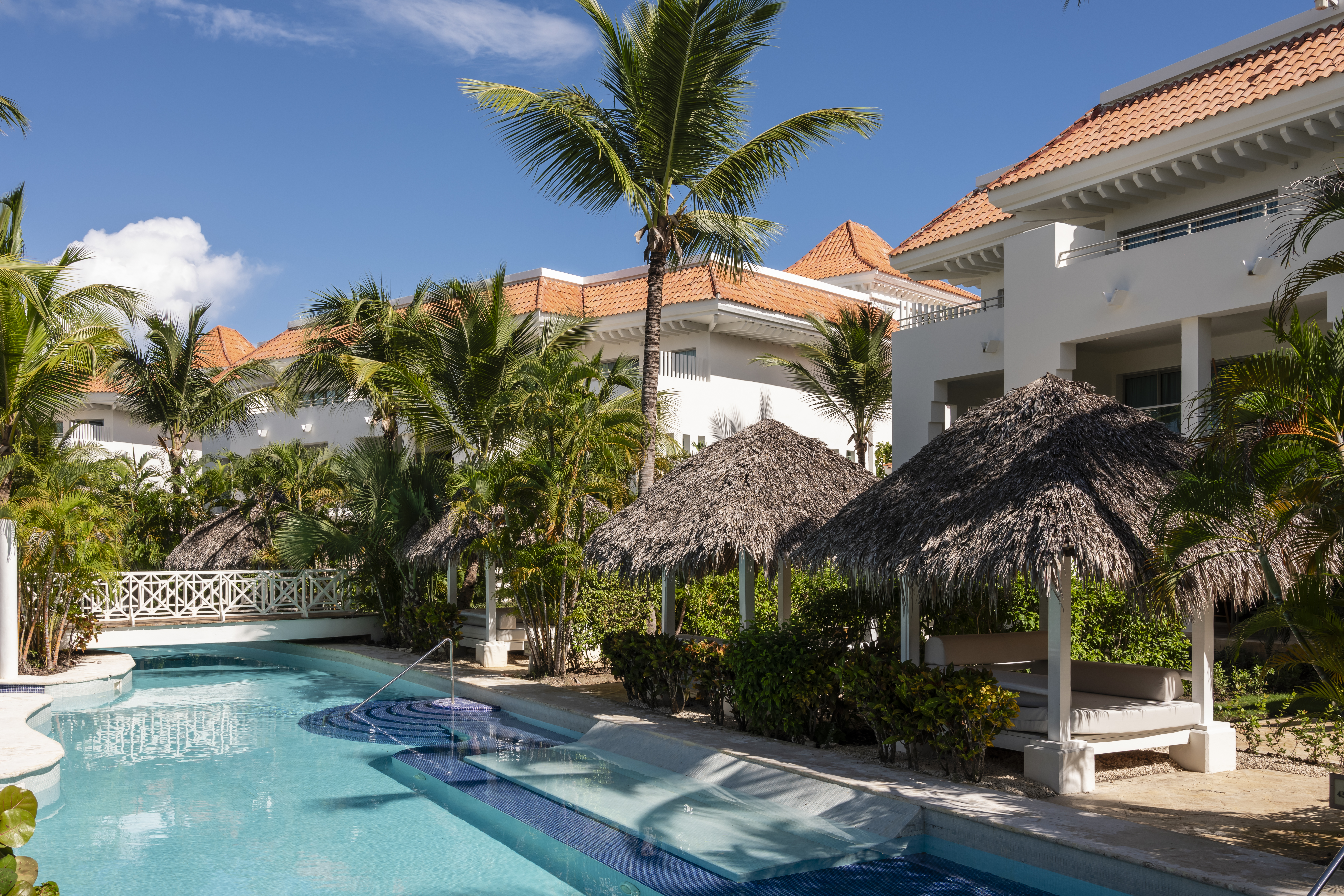Sunny tropical resort pool with white buildings, palm trees, and thatched cabanas.