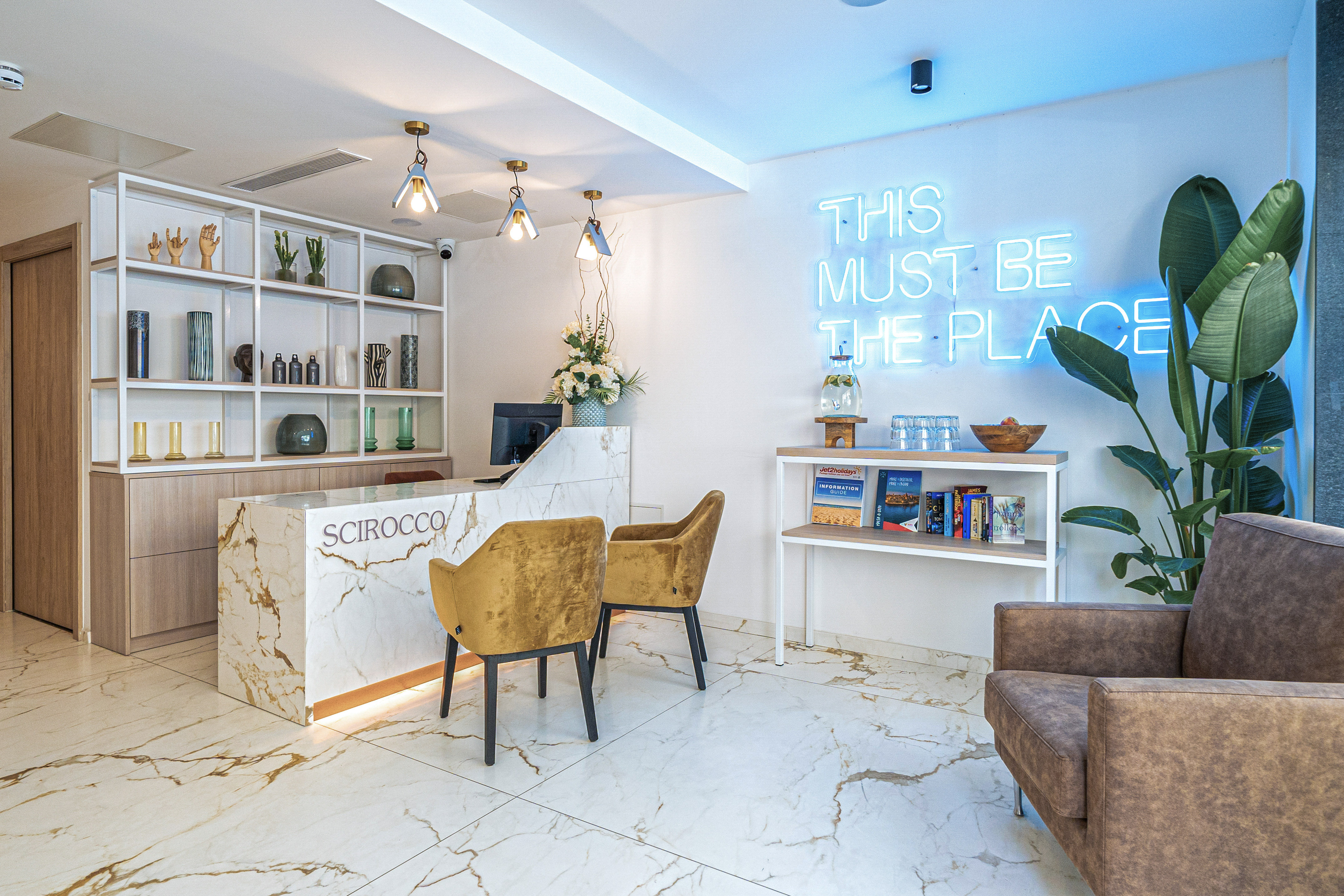 Scirocco hotel reception with marble desk, "THIS MUST BE THE PLACE" neon sign.