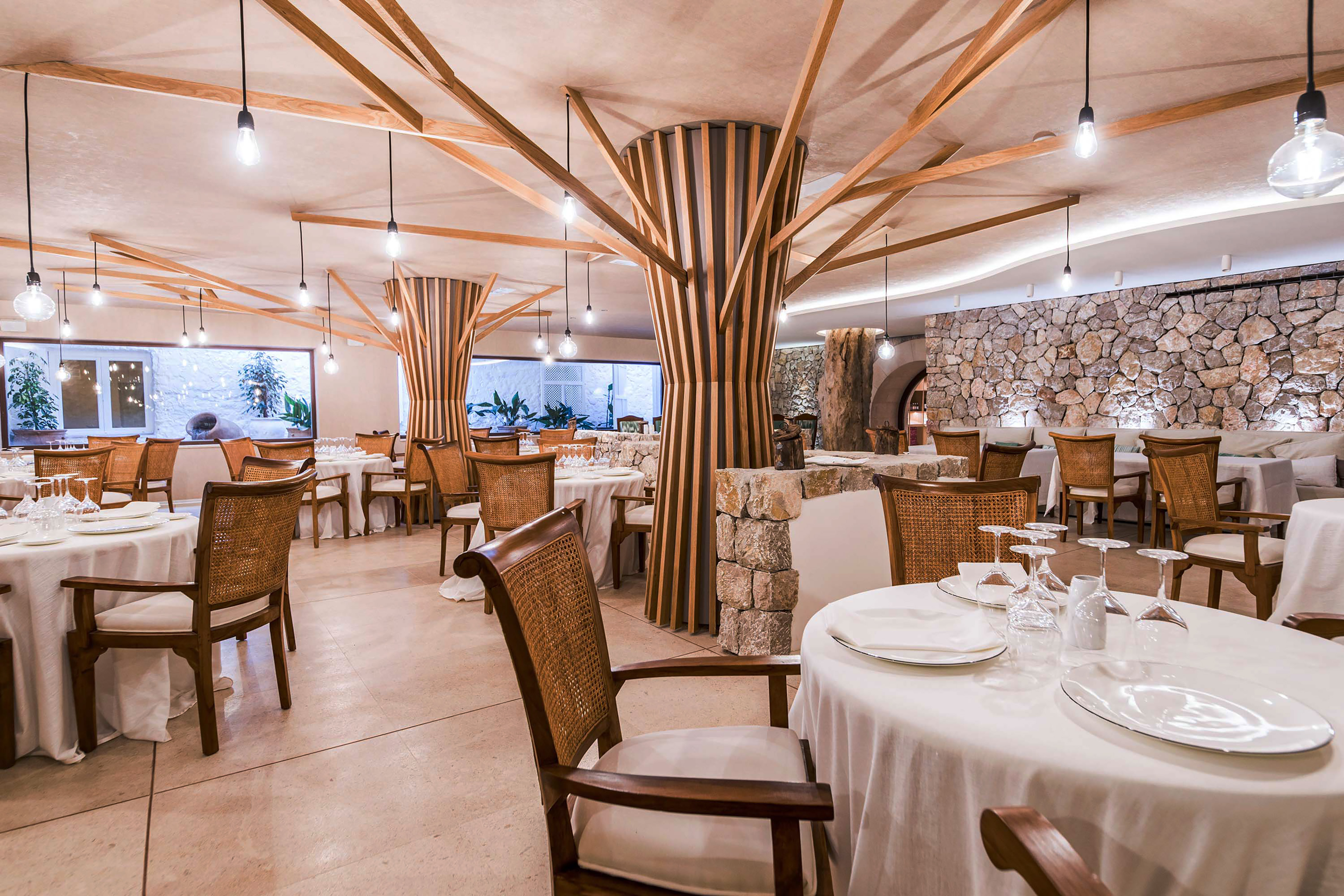 Modern restaurant interior with wooden tree columns, stone walls, and set tables.