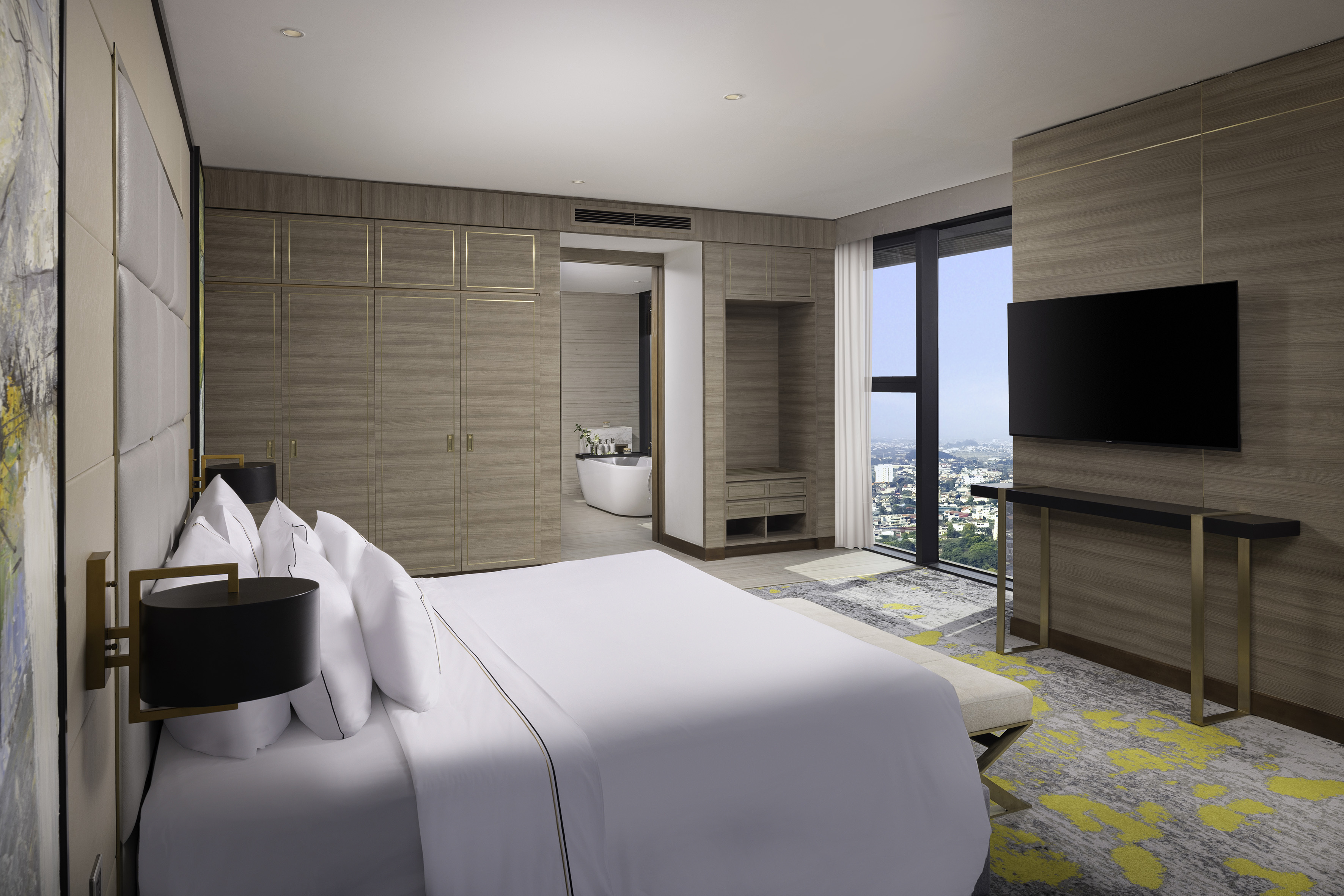 Modern hotel bedroom suite with city view, king bed, wood paneling, open bath.
