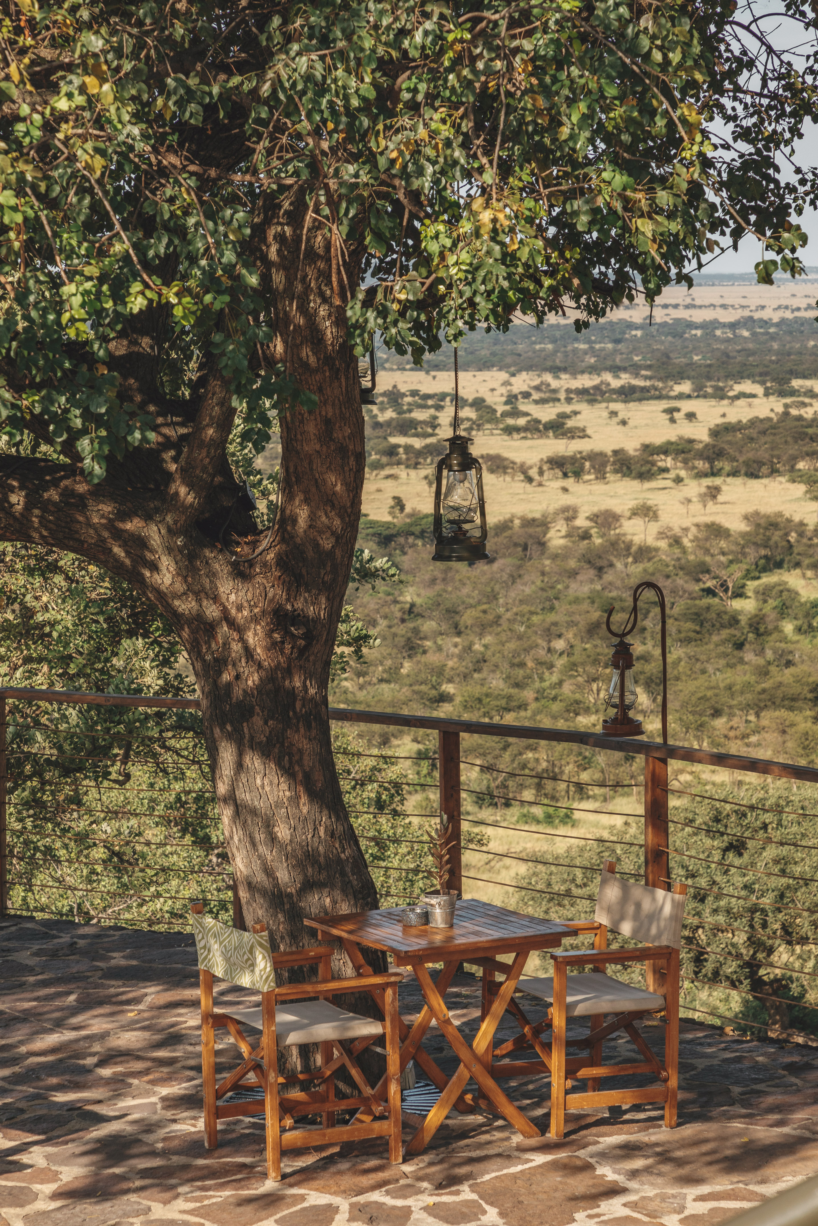 Patio with wooden table, chairs, lanterns, overlooking a vast African savanna.
