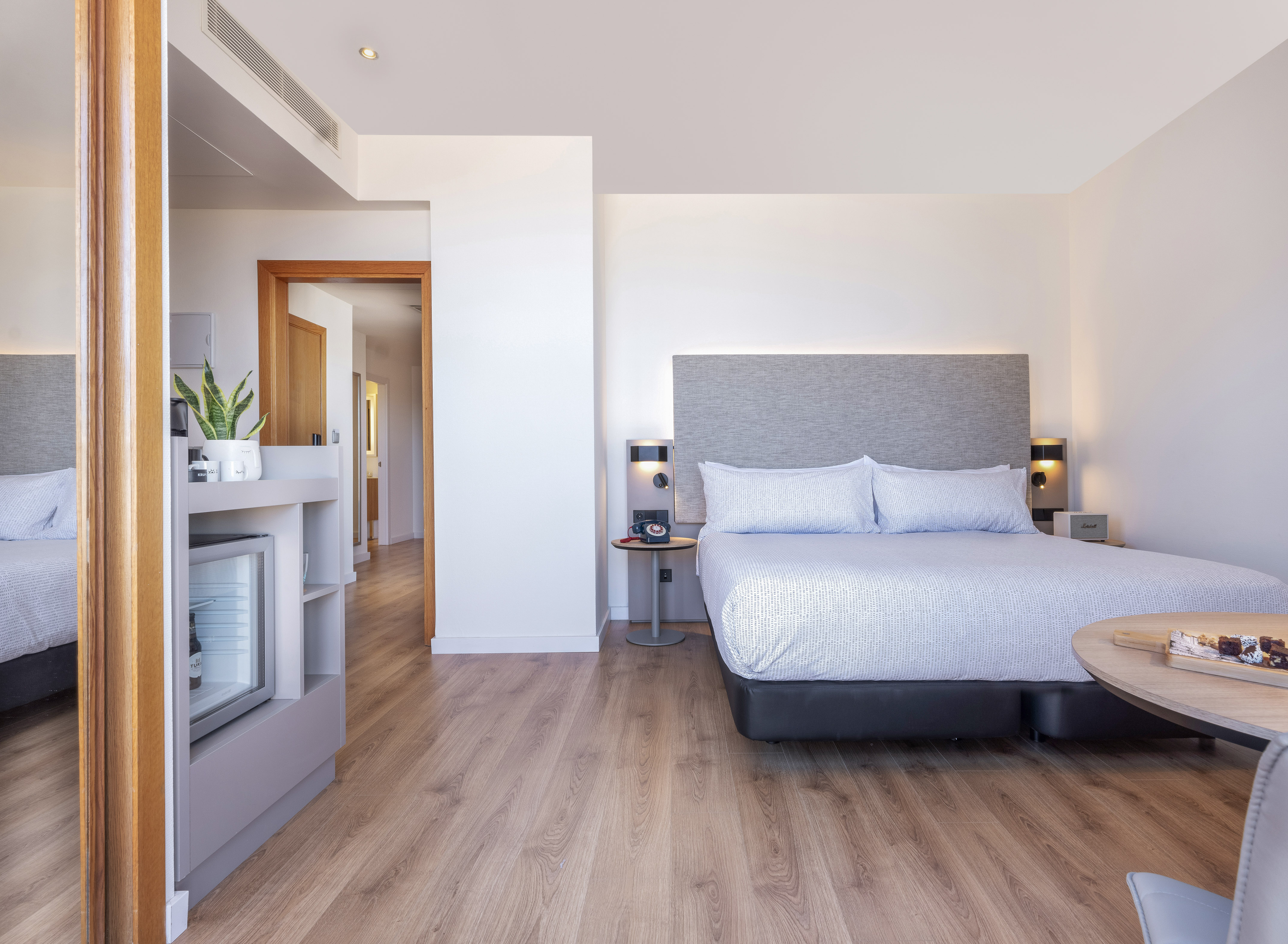 Modern hotel suite with bed, minibar, snacks, and wooden floor.