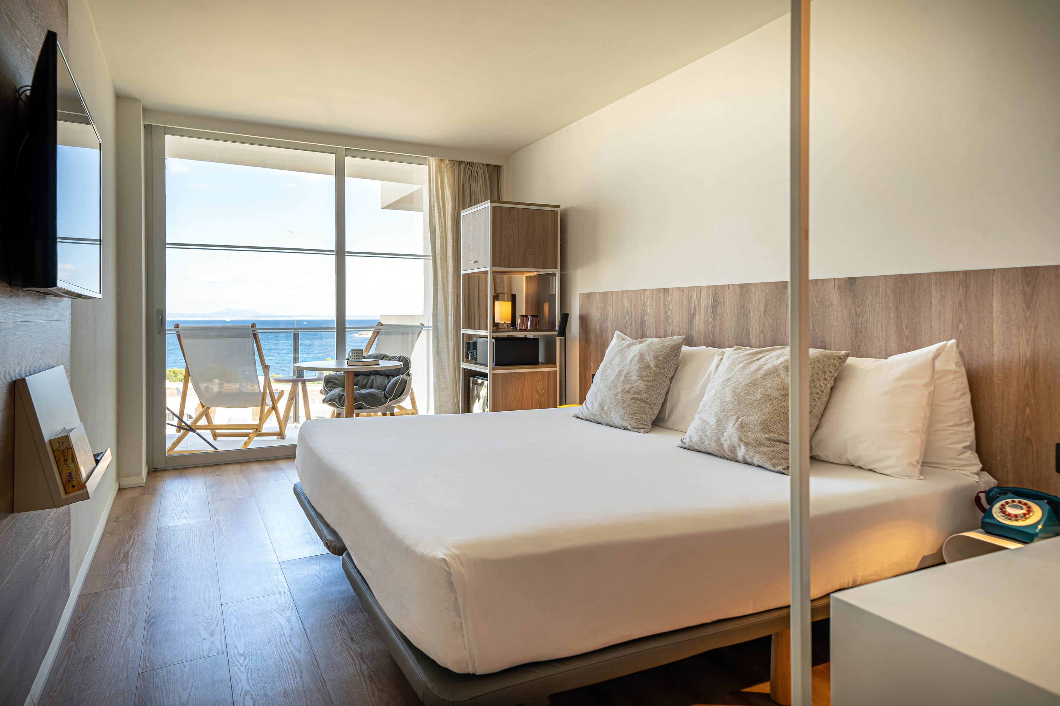 Modern hotel room with sea view balcony, white bed, and wooden floor.