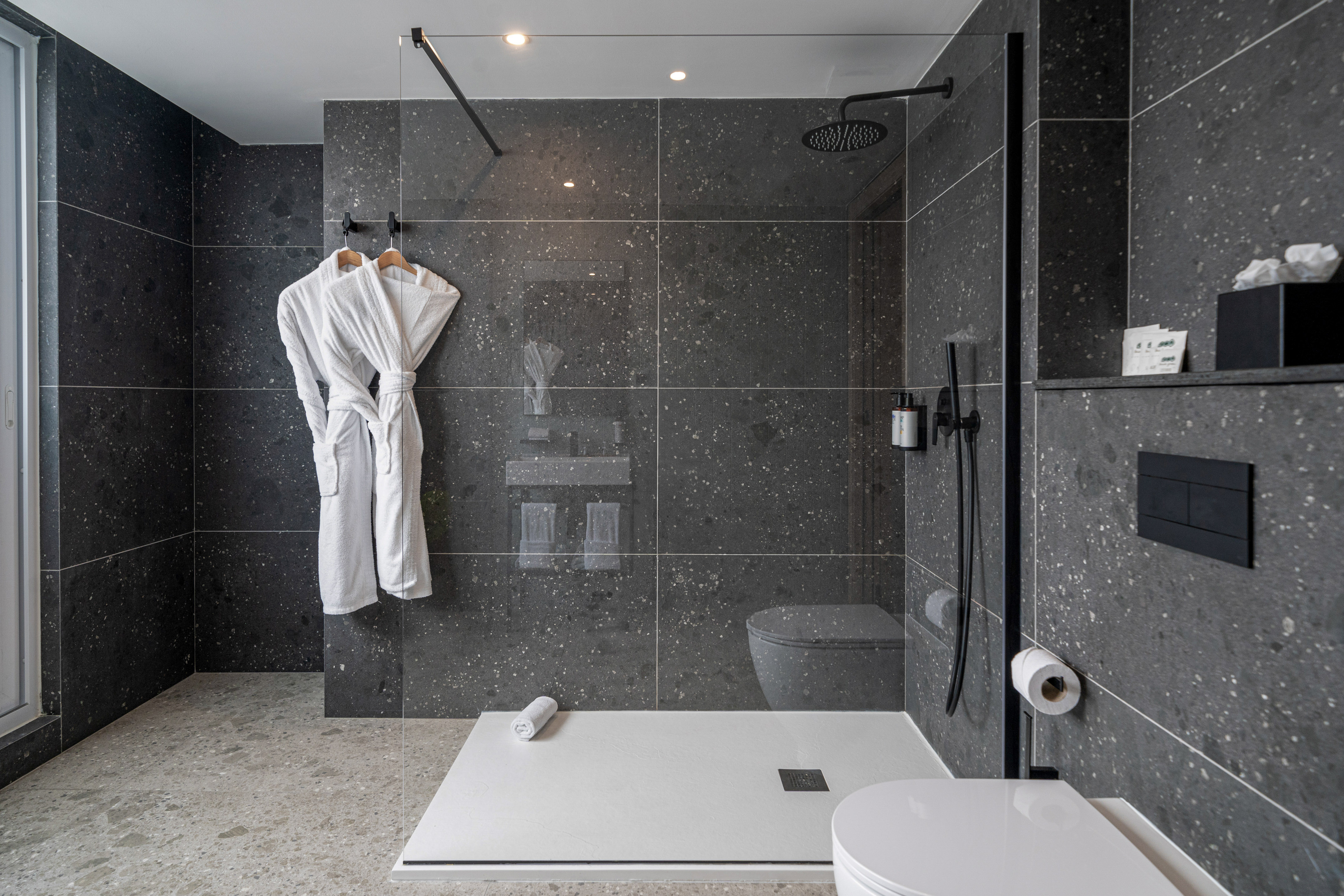 Modern bathroom with dark terrazzo tiles, glass shower, and white bathrobes.