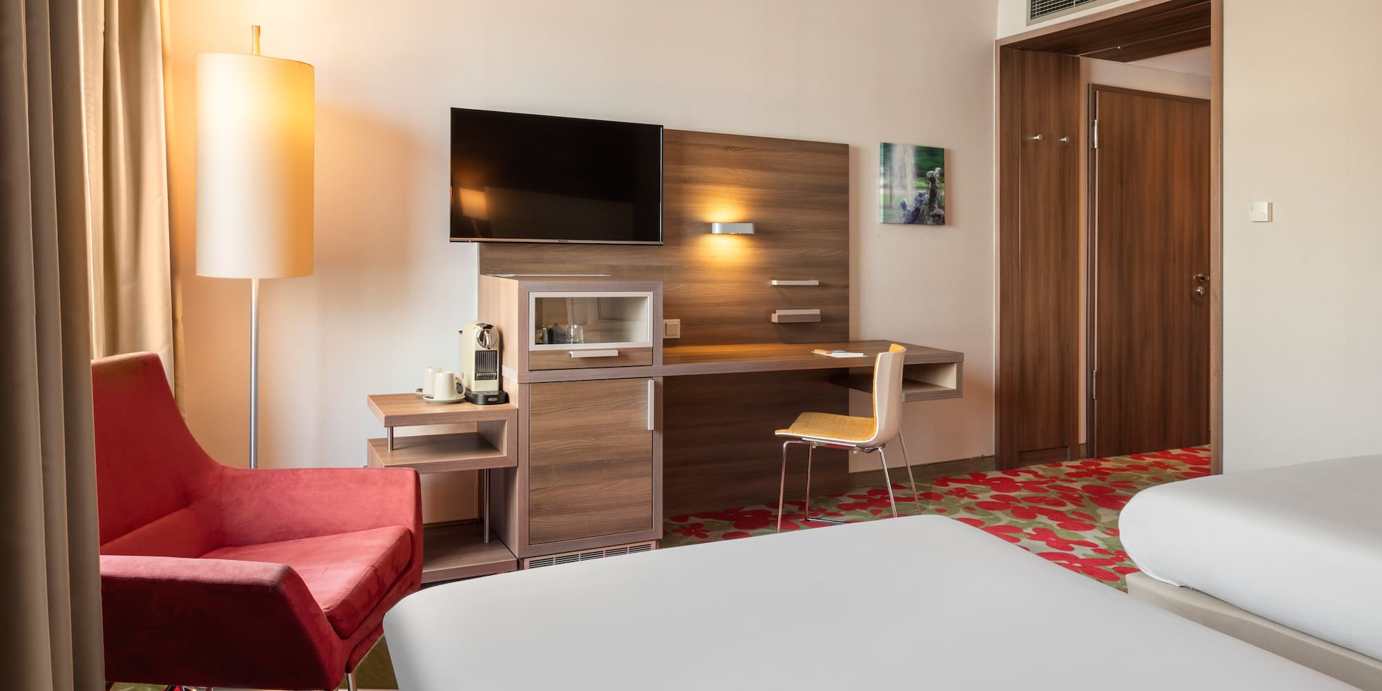 Modern hotel room with two beds, red armchair, desk, TV, and coffee machine.