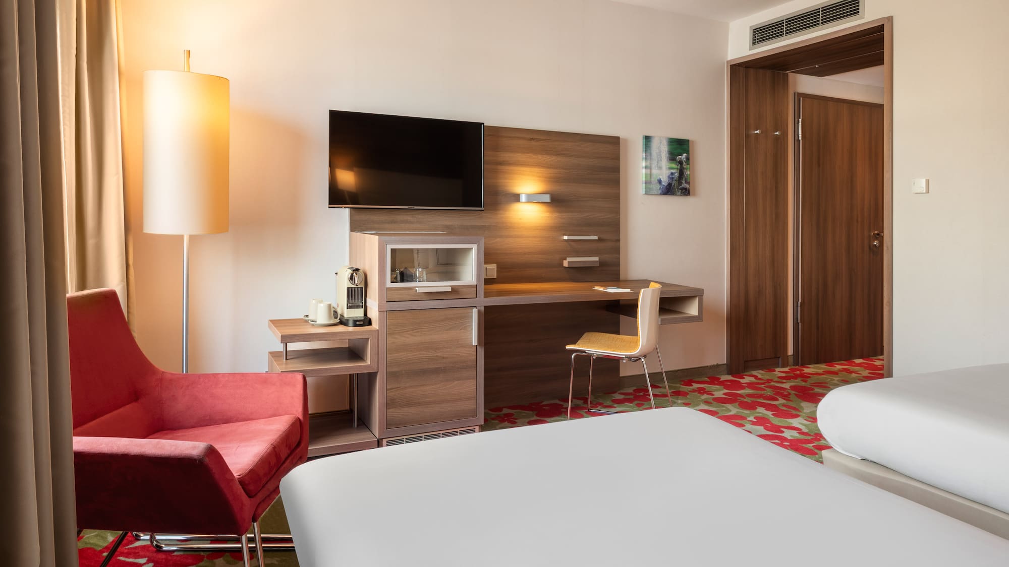 Modern hotel room with two beds, red armchair, desk, TV, and coffee machine.