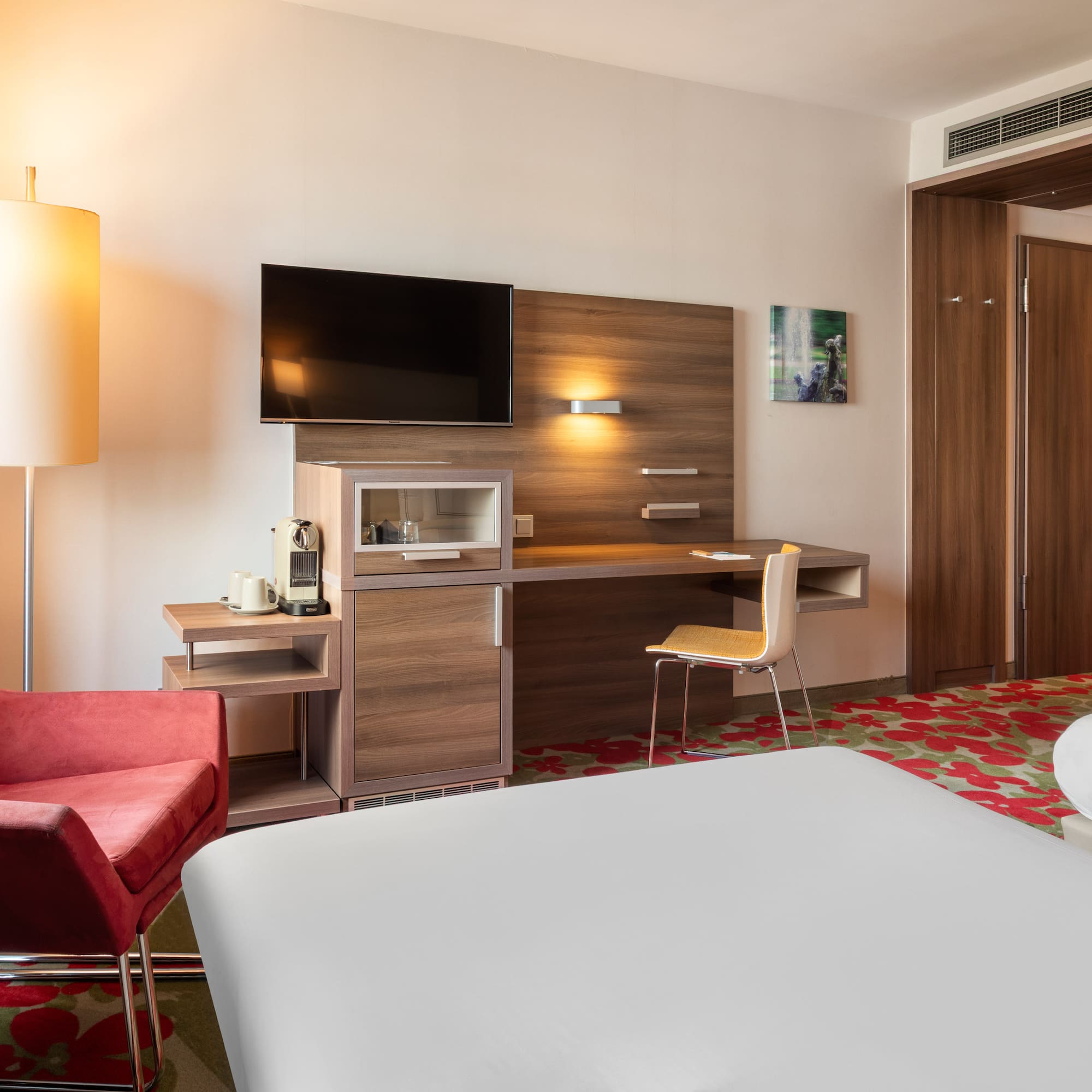 Modern hotel room with two beds, red armchair, desk, TV, and coffee machine.