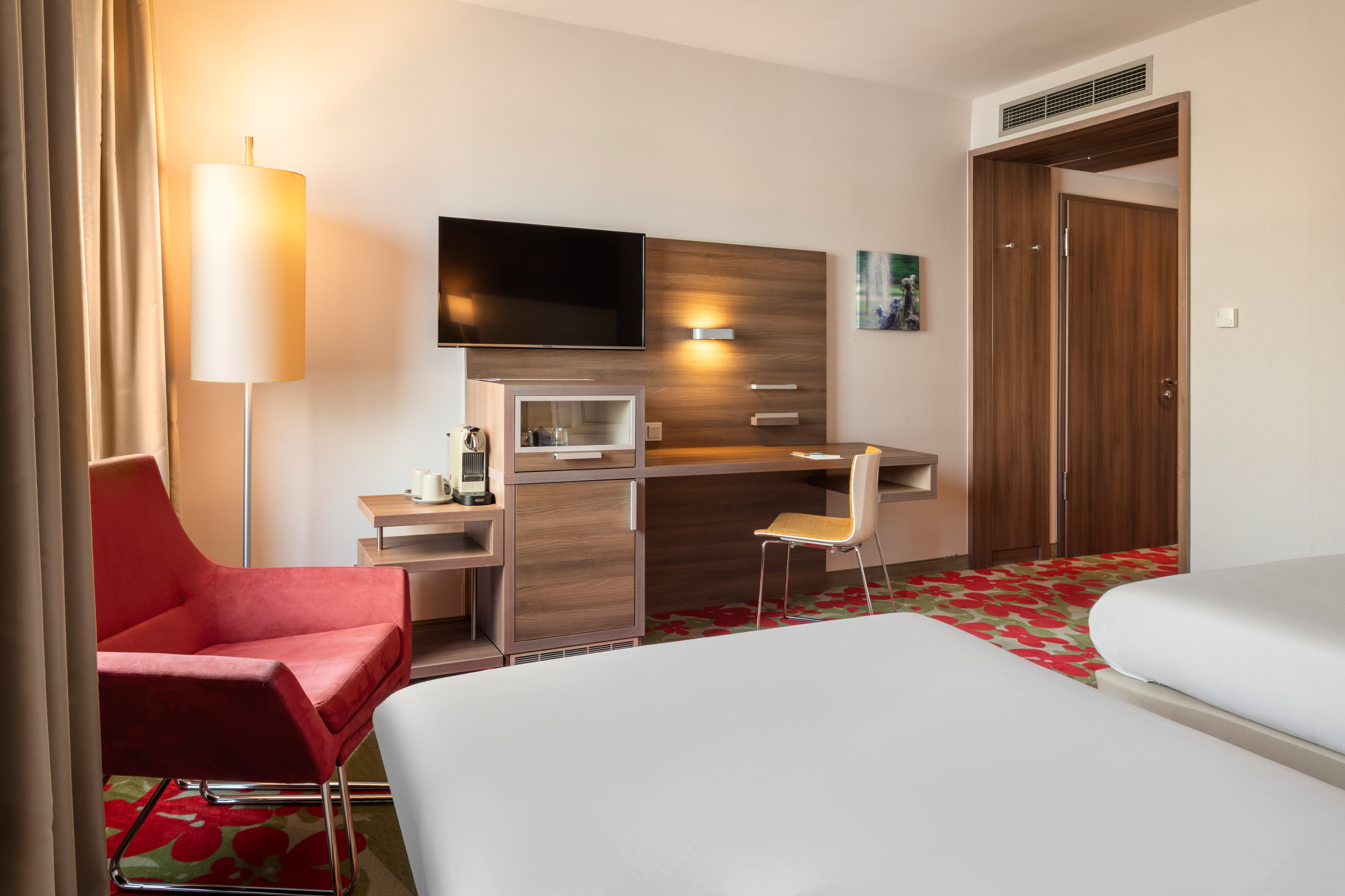 Modern hotel room with two beds, red armchair, desk, TV, and coffee machine.