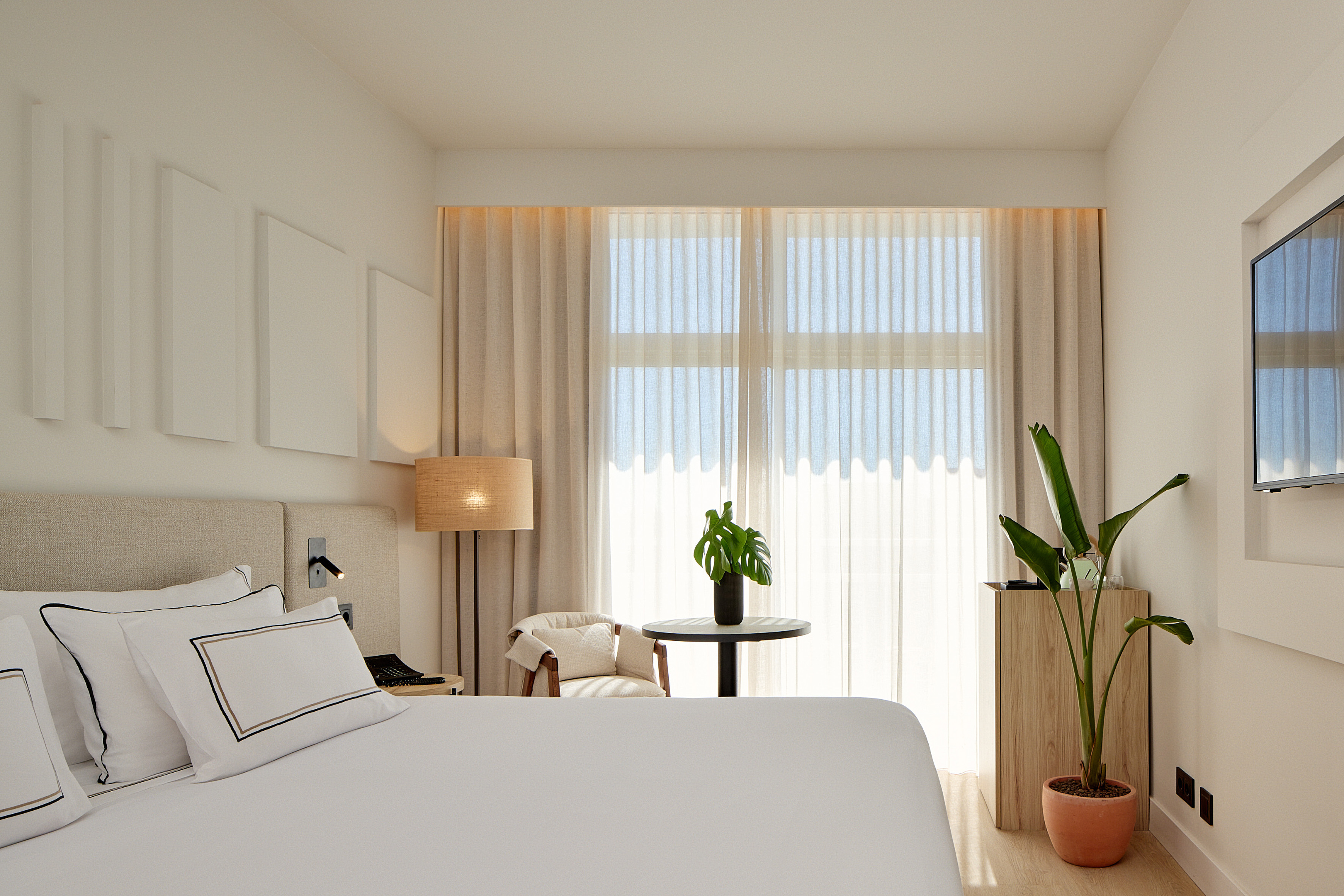 Minimalist hotel bedroom featuring white bedding, a bright window, and houseplants.