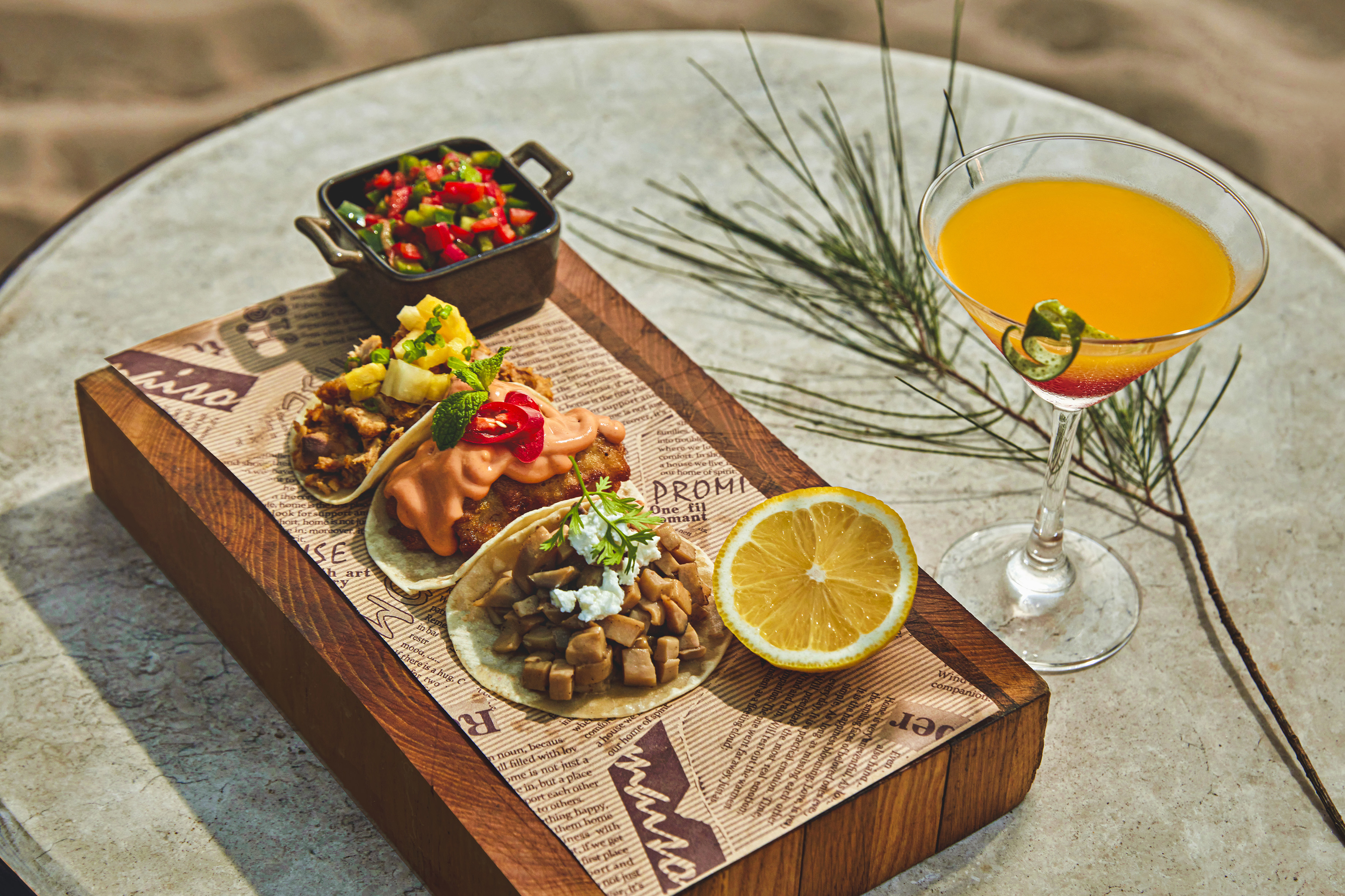 Three tacos, fresh salsa, lemon, and a tropical orange cocktail on wooden board.