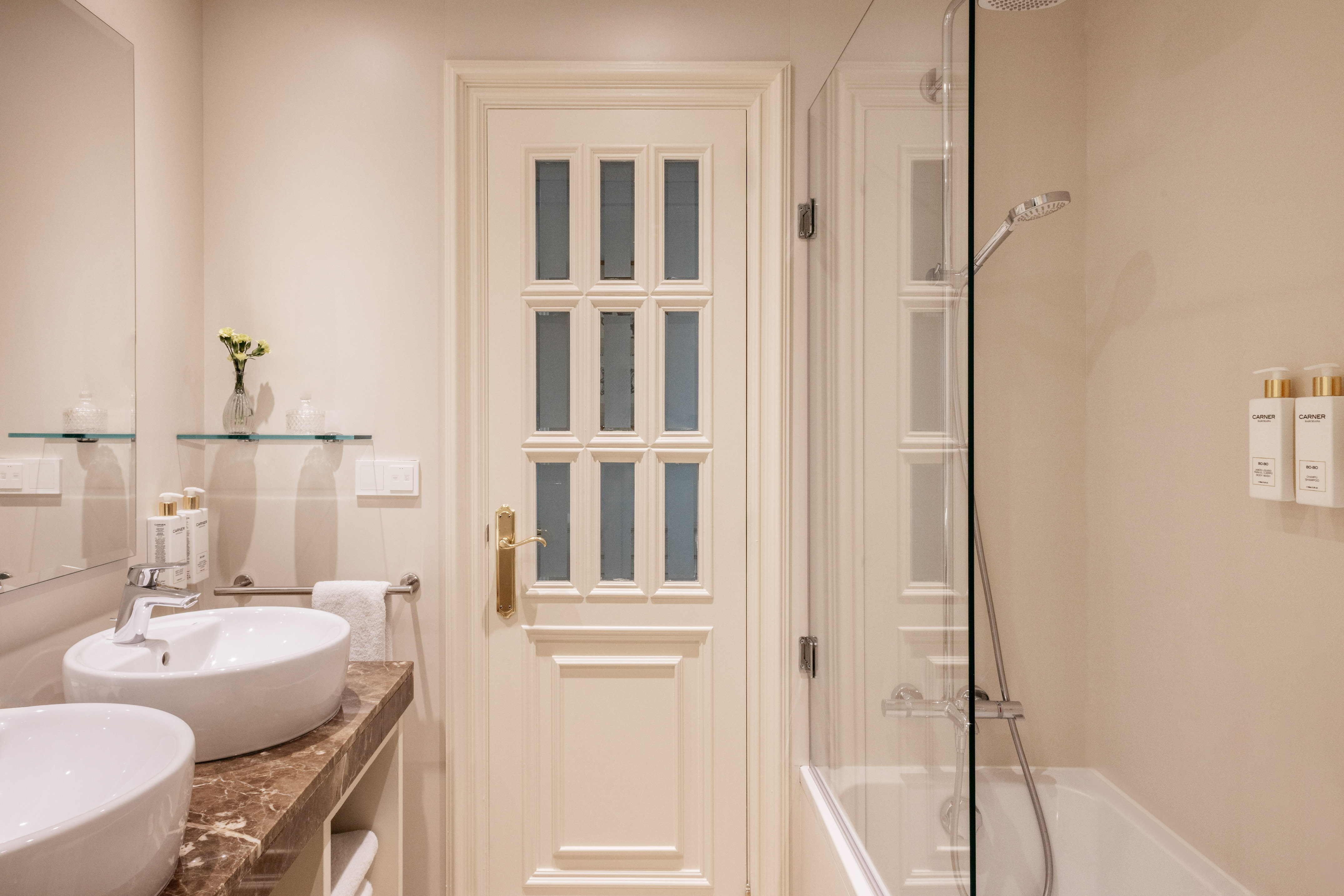 Hotel bathroom with double vessel sinks, marble vanity, glass shower, paneled door.