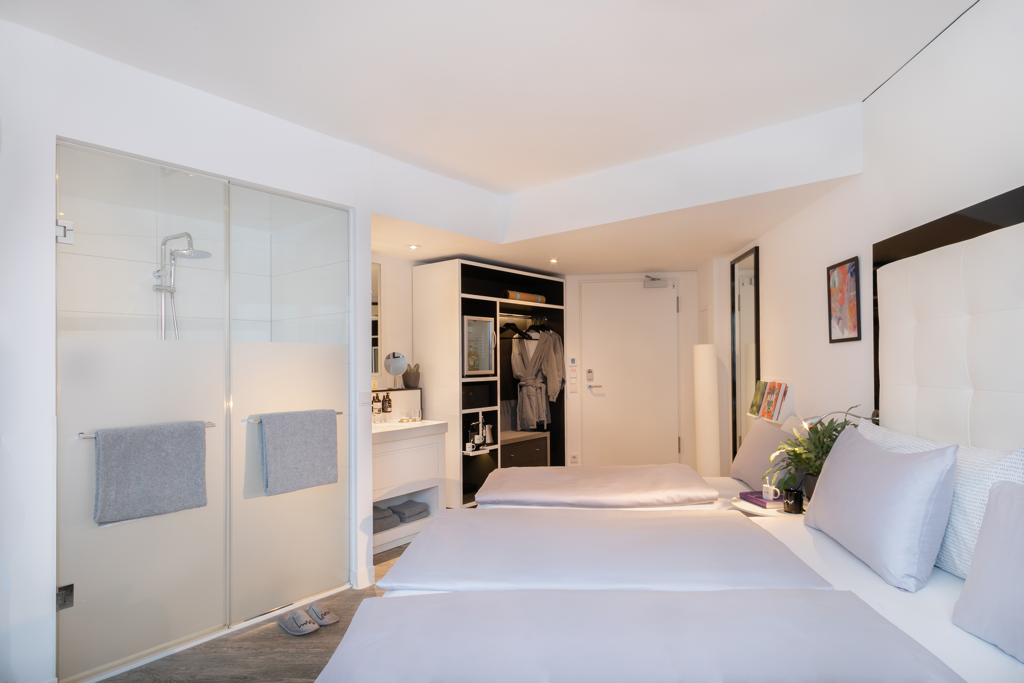 Bright hotel room with two beds, modern glass shower, and integrated wardrobe.