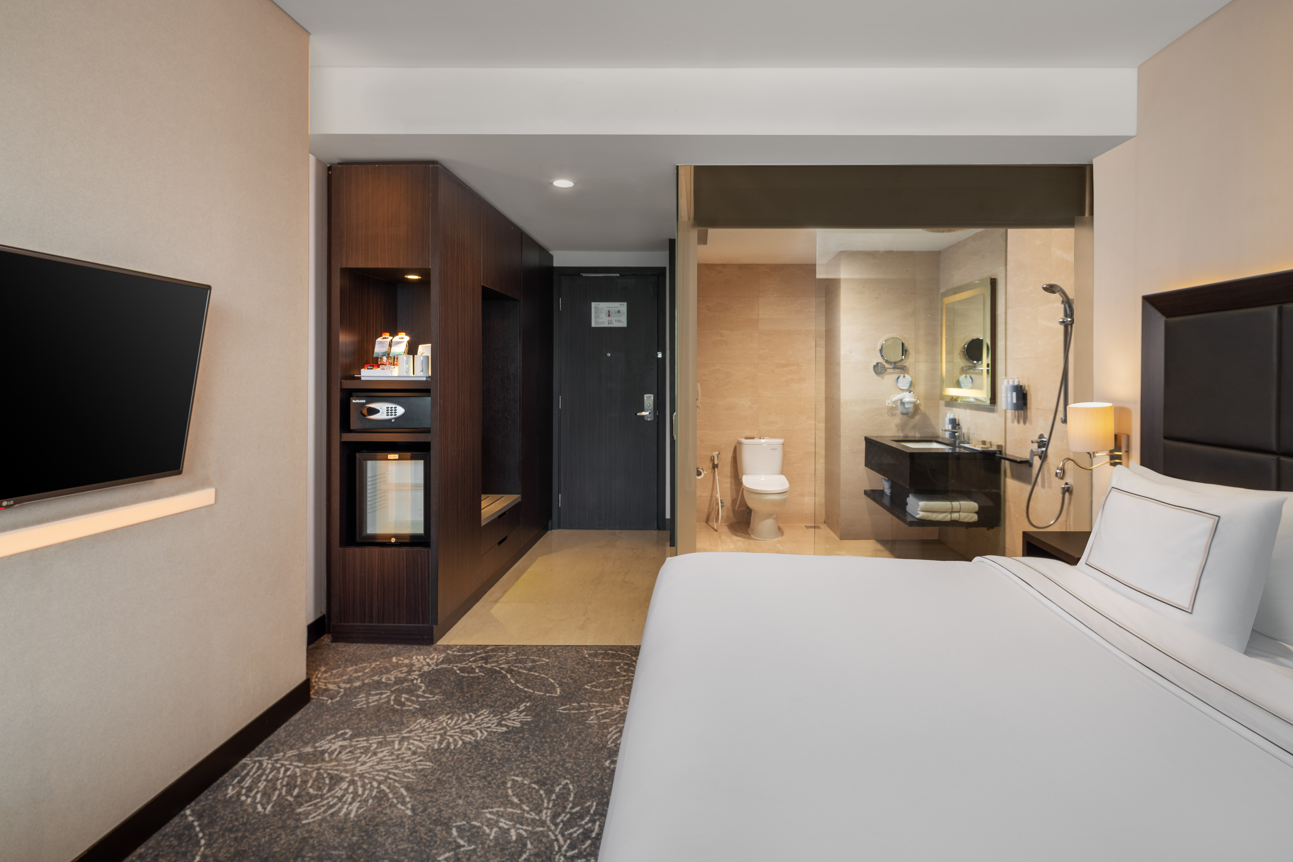 Modern hotel room with bed, TV, dark storage unit, and glass bathroom.