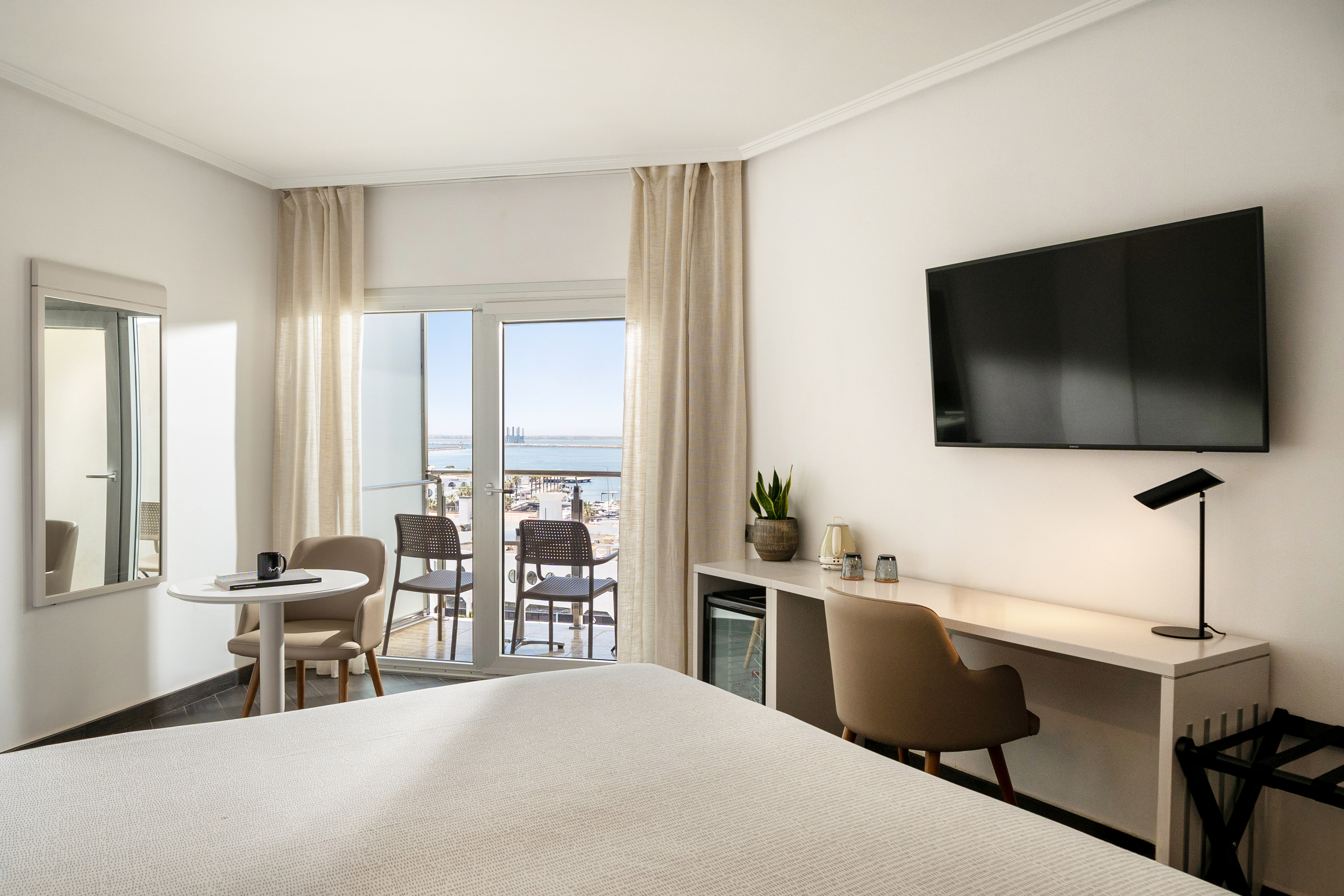 Bright hotel bedroom with sea view balcony, workspace, and modern furnishings.